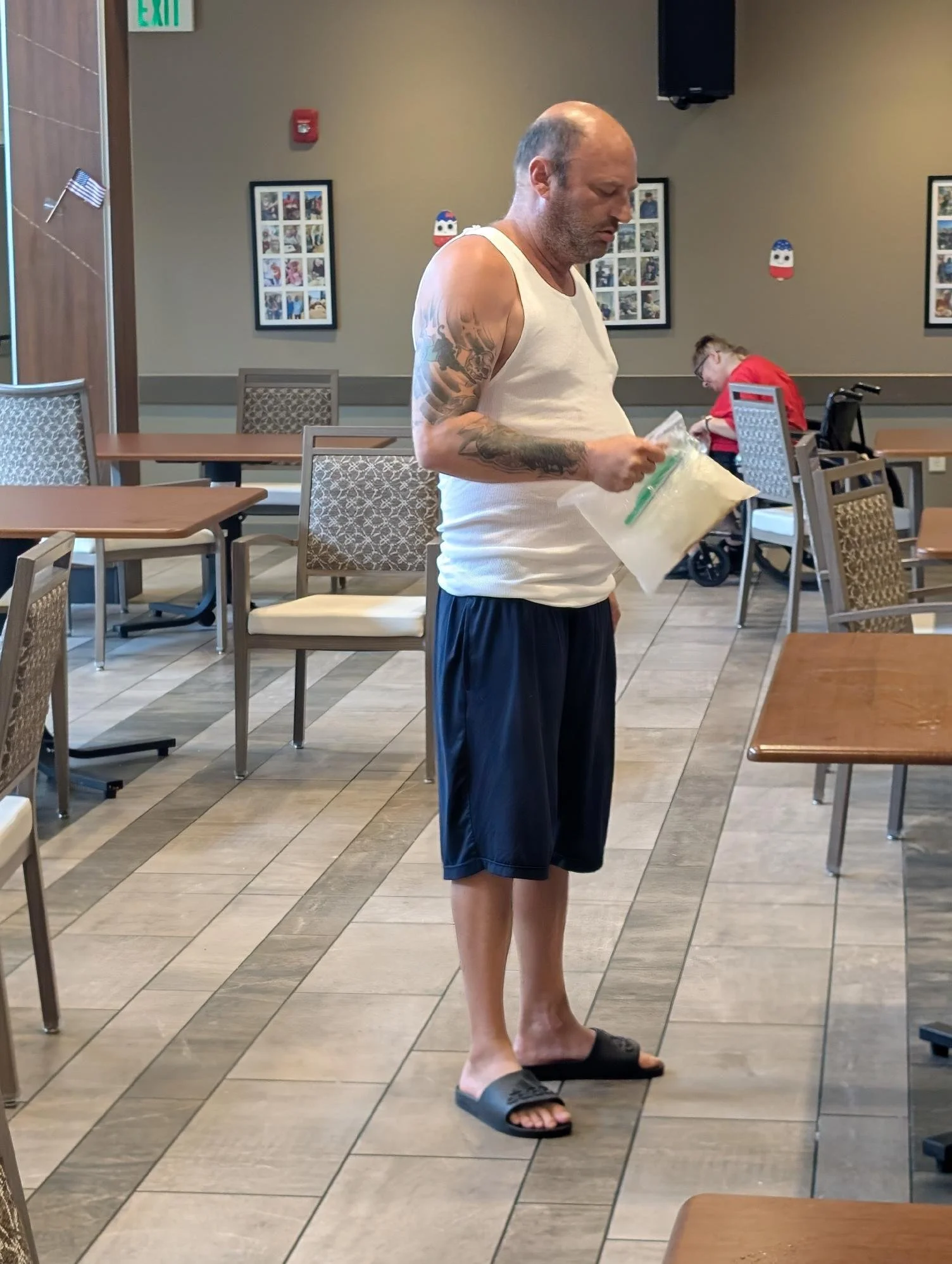 A man standing inside a restaurant wearing a white tank top, black shorts, and slippers, holding a plastic bag with his left hand, with a woman in the background sitting at a table in a red shirt and a wheelchair.