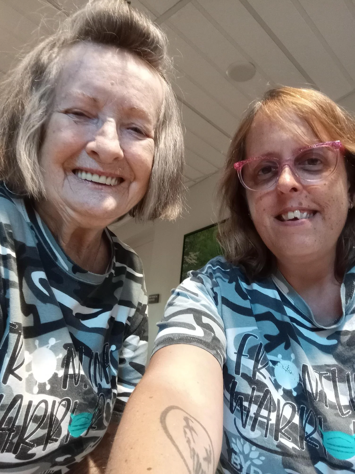 Two women taking a selfie together, wearing matching shirts with similar black, white, gray, and blue patterns, smiling in an indoor setting.