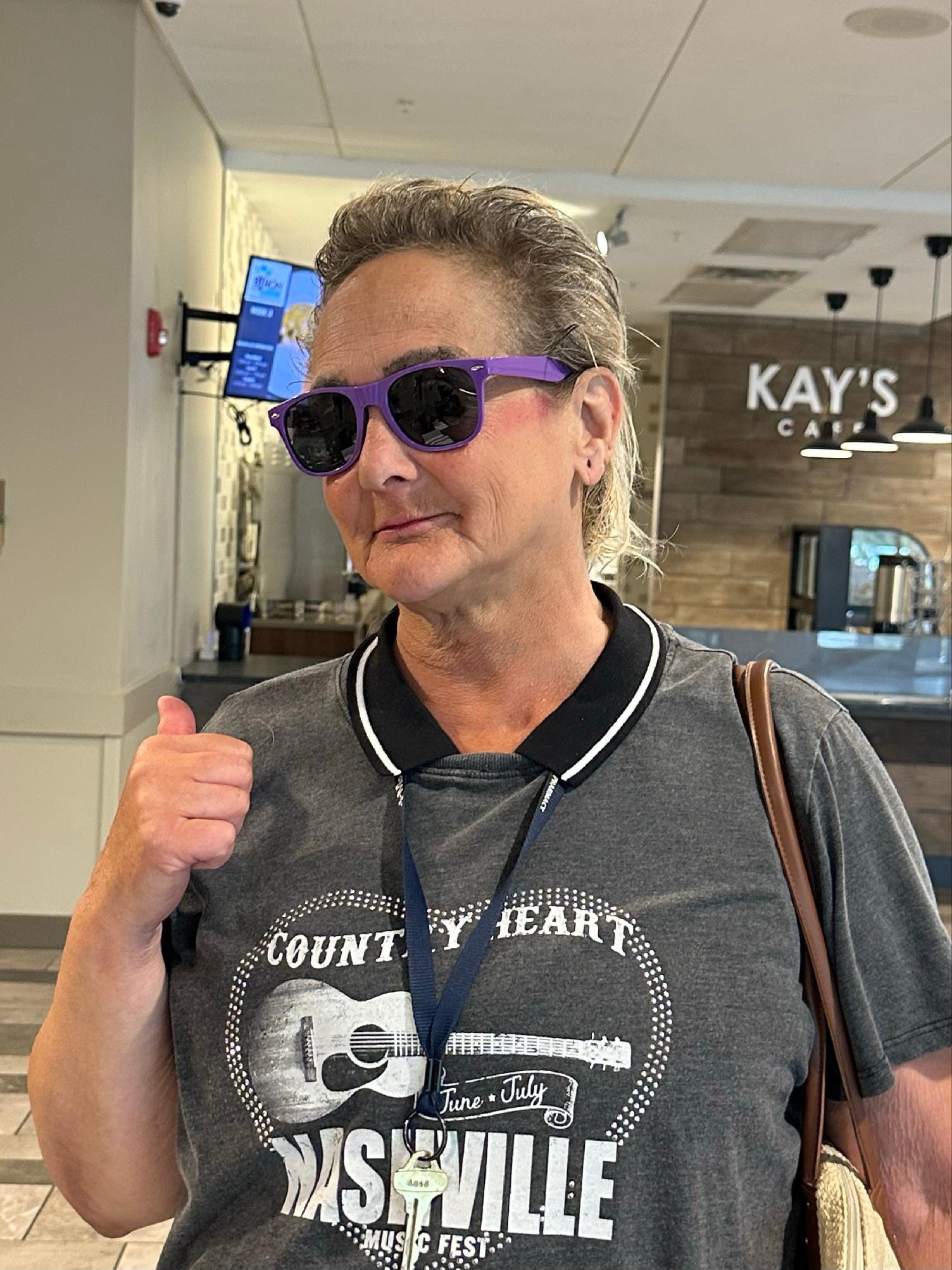 A woman with blonde hair wearing purple sunglasses and a gray T-shirt with Nashville music festival graphics, giving a thumbs up inside a cafe with a sign that reads 'KAY'S CAFE' in the background.