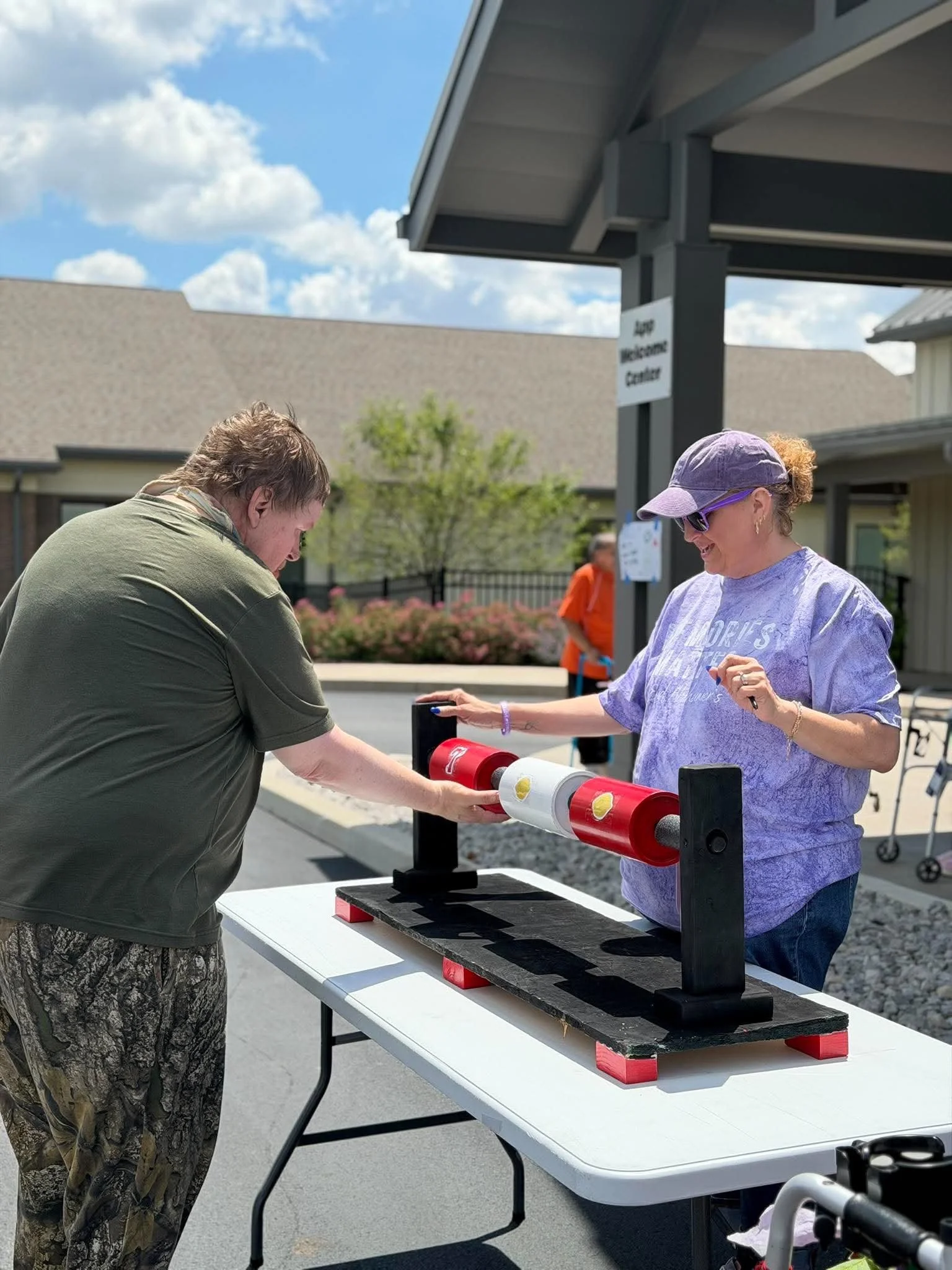 A woman and a man participate in an outdoor event, placing yellow tennis balls into a rolling tube contraption on a white table. The woman is wearing a purple cap, sunglasses, a lavender t-shirt, and is smiling. The man has a green shirt and camoufla