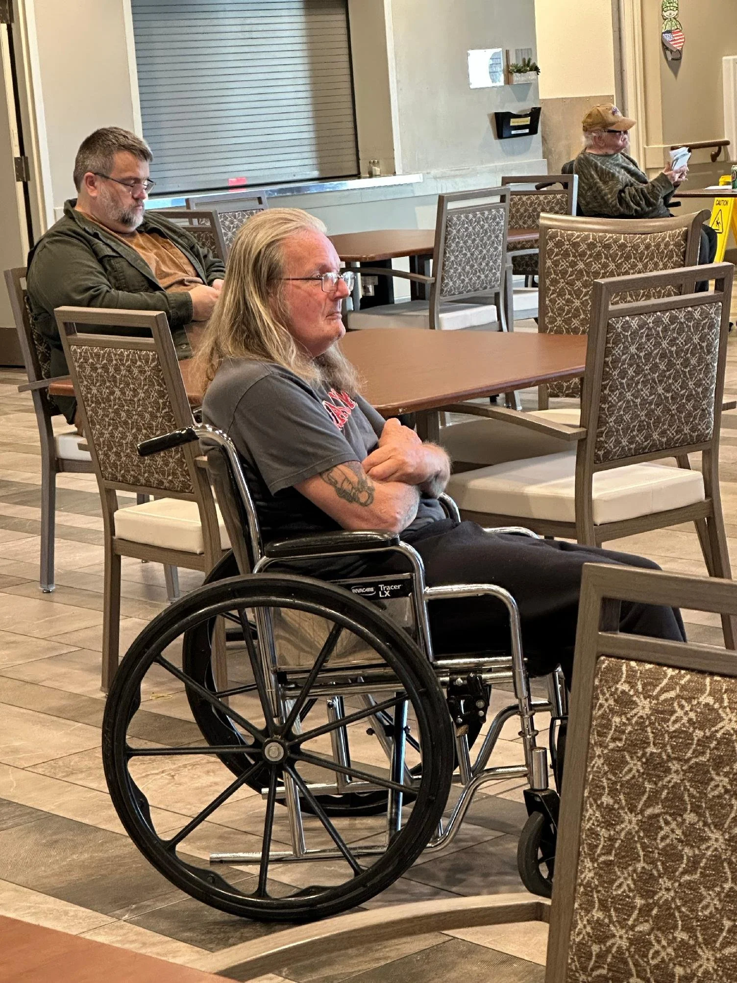 Three older adults seated apart in a waiting area, two in chairs and one in a wheelchair, all looking at their phones.