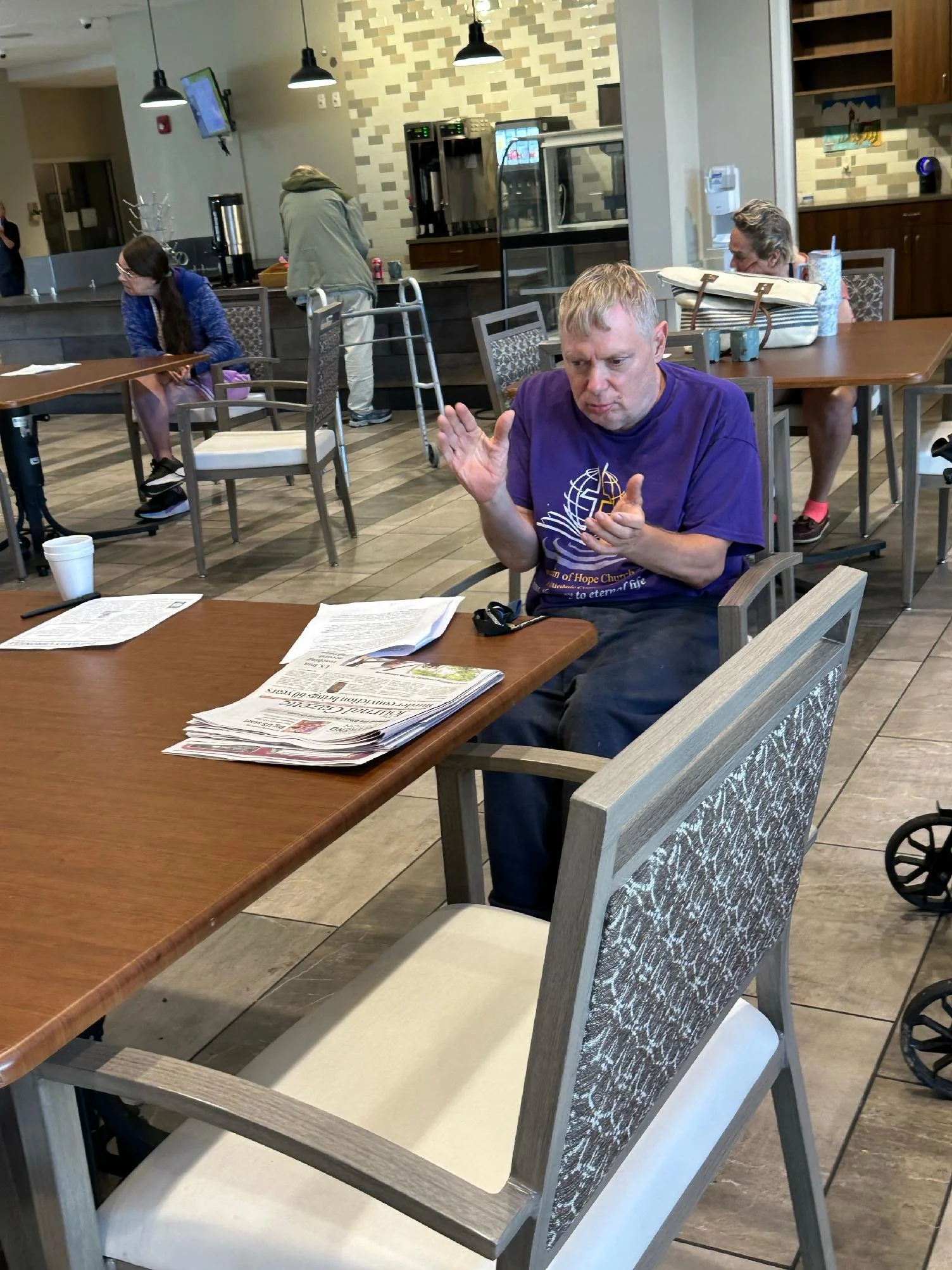 People seated at a dining area in a modern coffee shop or restaurant. A man in a purple shirt is front and center, with reading materials on the table. Behind him, a woman with glasses and a blue sweater, a woman with a large tote bag, and a person w