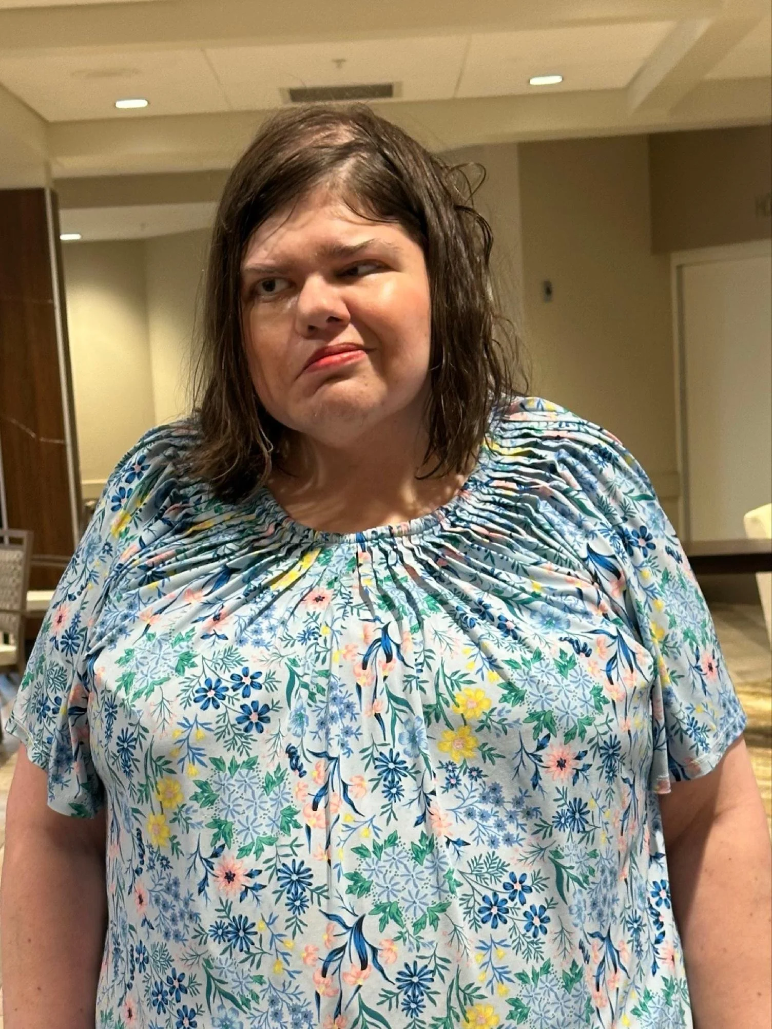 Woman with shoulder-length brown hair wearing a colorful floral-patterned blouse, standing indoors in a well-lit room.