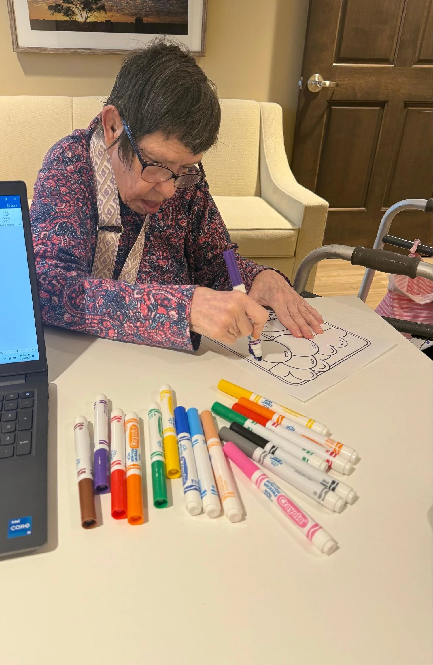 An elderly woman with short gray hair, glasses, and a patterned shirt is sitting at a table, coloring a picture with a purple marker. Multiple colorful markers are scattered on the table in front of her. In the background, there is a beige sofa, a framed picture of a landscape hanging on the wall, and a partially open wooden door.