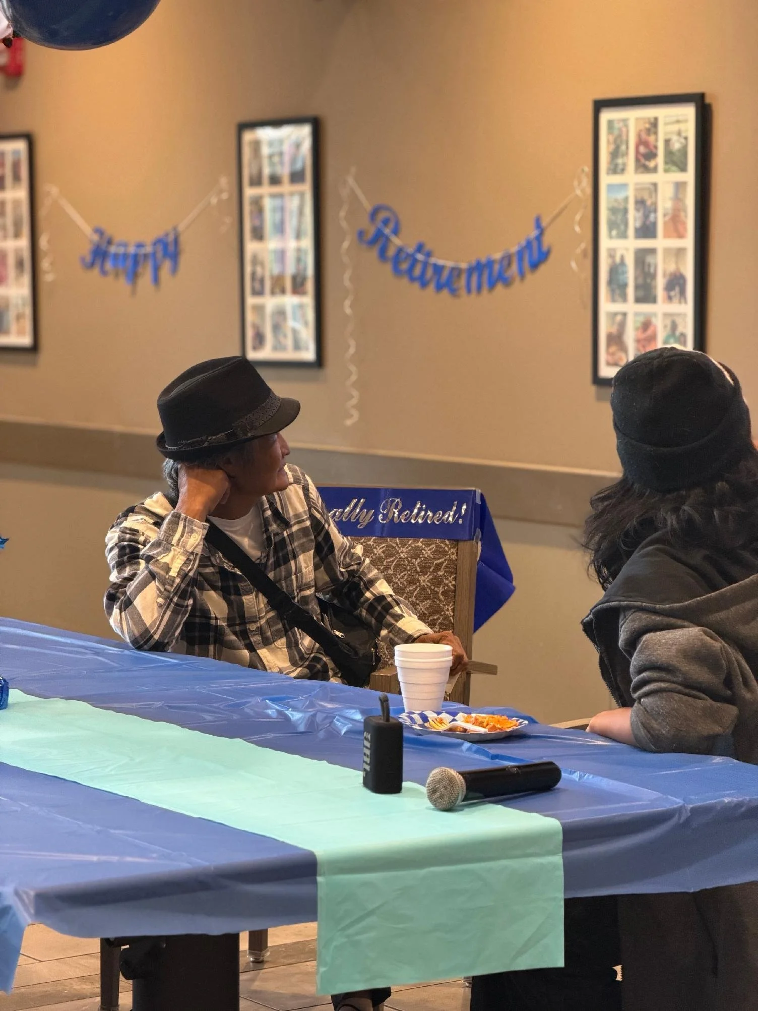 People sitting at a table decorated with teal and navy blue tablecloths, with a microphone, snacks, and a white cup on the table in a decorated indoor setting with framed pictures on the wall and a blue banner that reads "Really Retired!"