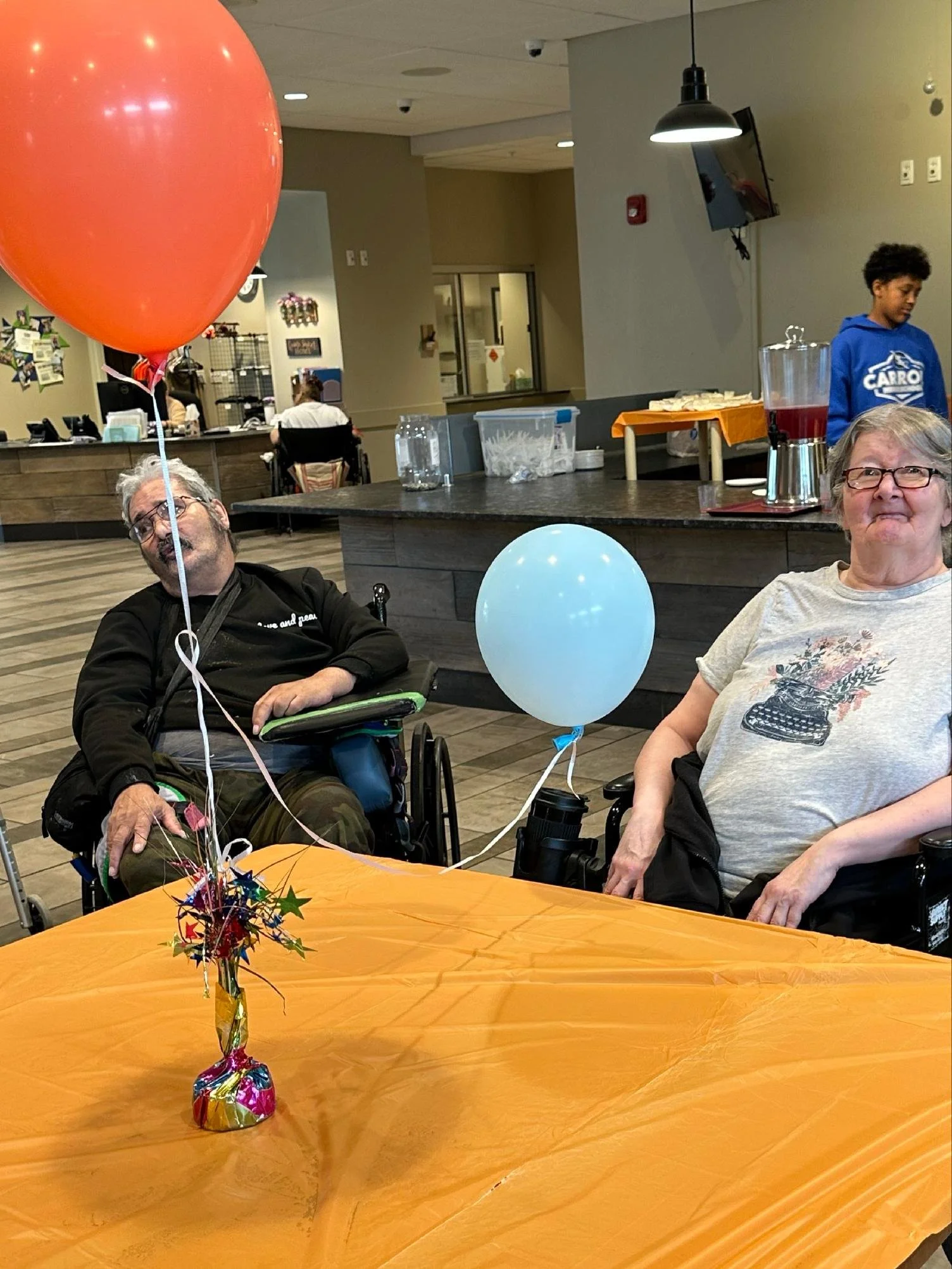 Two elderly individuals sitting at a table decorated with a gold-colored tablecloth and a small colorful party centerpiece. One is holding balloons, and both are smiling. There is a large red balloon in the foreground. The scene appears to be in a co