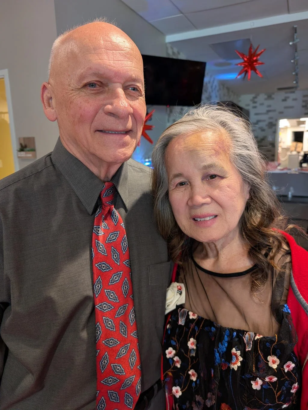An elderly man and woman are smiling and posing for the photo indoors, with festive decorations like red star-shaped ornaments hanging from the ceiling in the background.