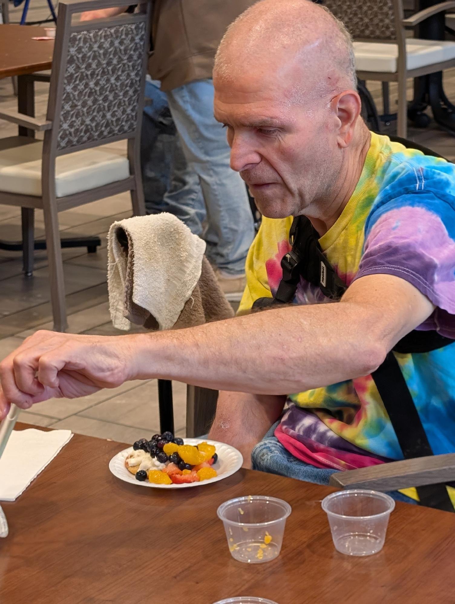 Man with a shaved head and tie-dye shirt sitting at a table with a plate of fruit salad, holding a towel, in a casual dining setting.
