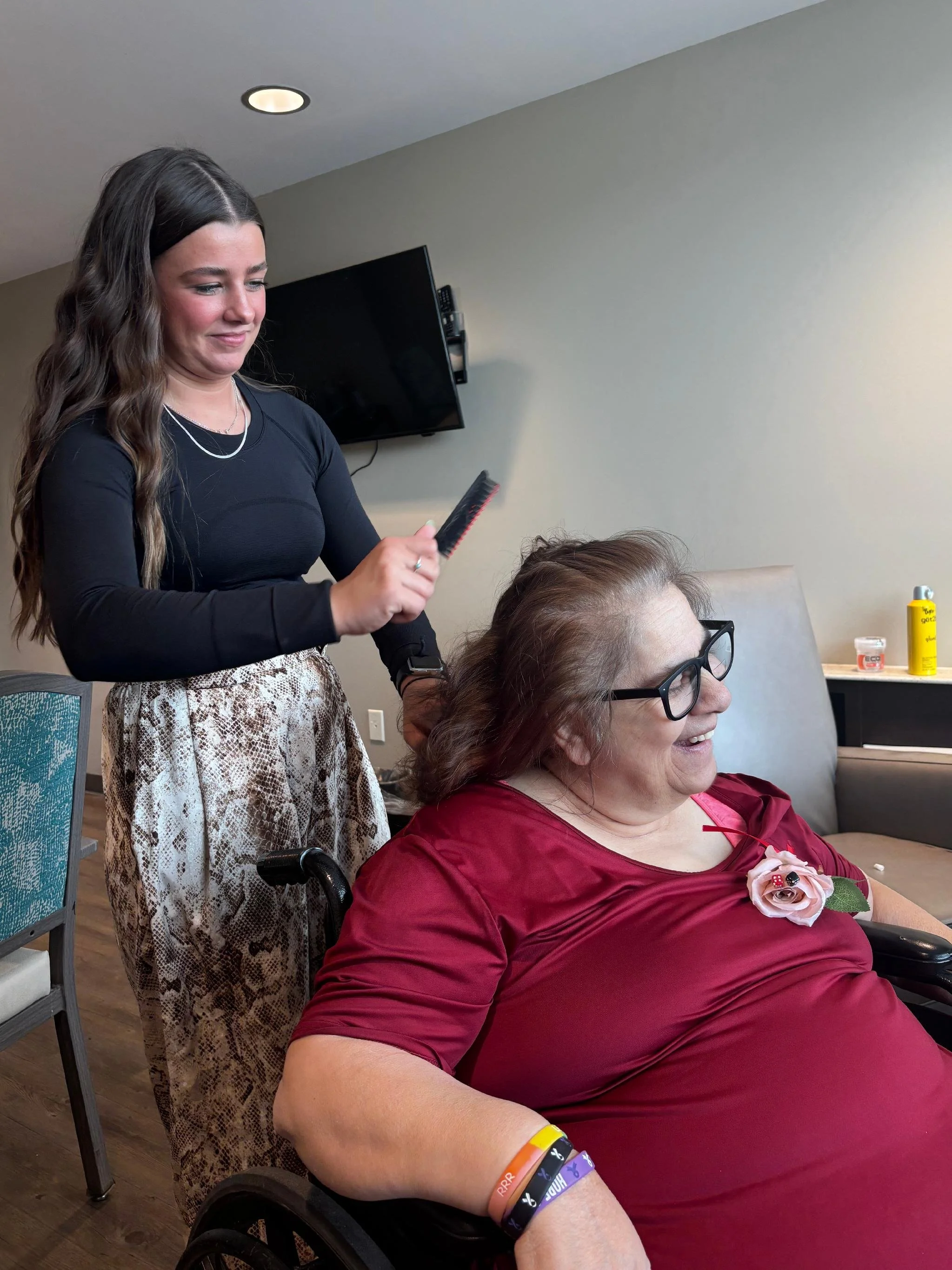 Young woman with long hair styling the hair of an older woman sitting in a wheelchair, both smiling; the older woman wears glasses, a red top, and a flower pin on her shirt.