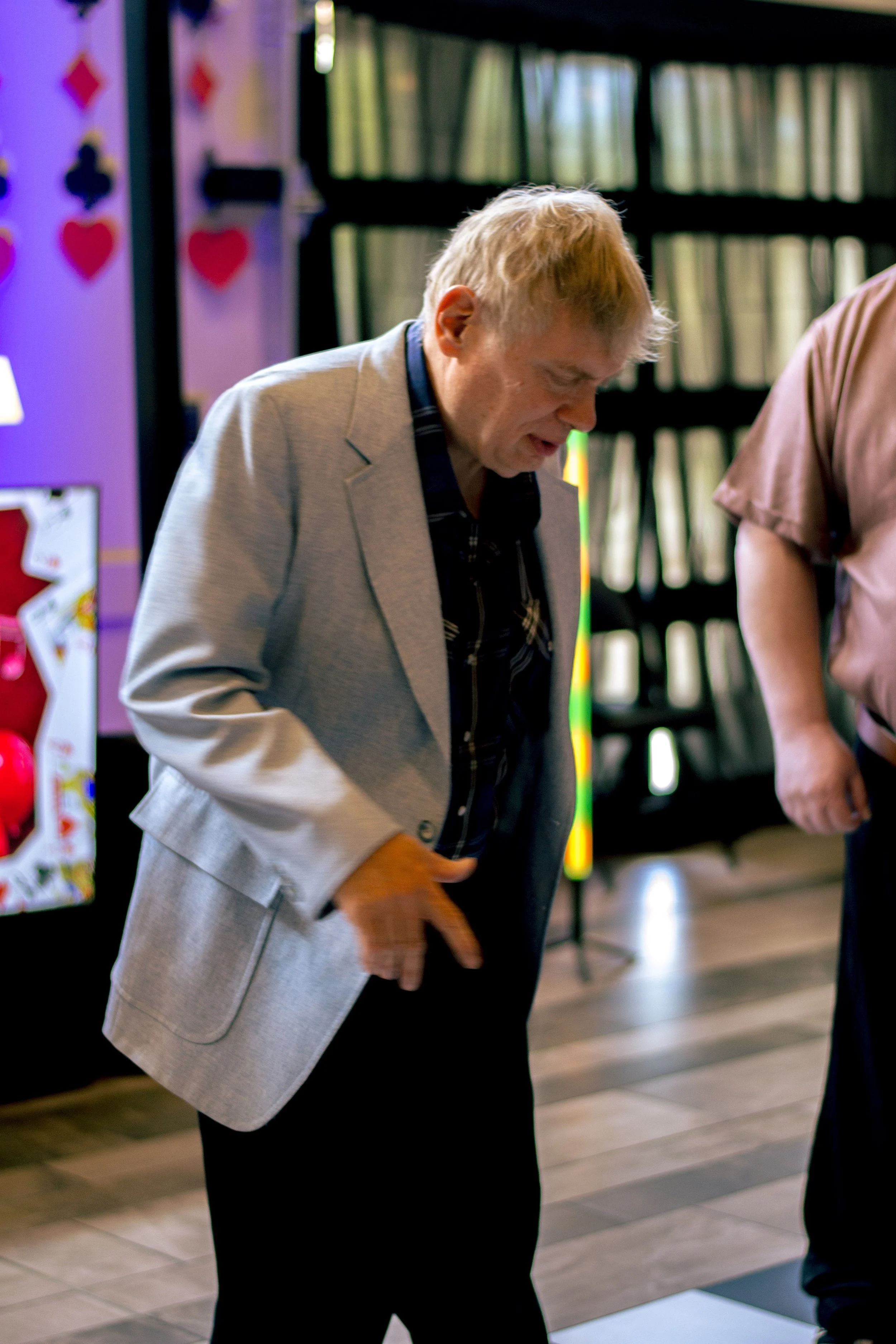 A man with blond hair wearing a gray blazer and dark shirt bowing with a smile in an indoor setting, possibly at a social event.