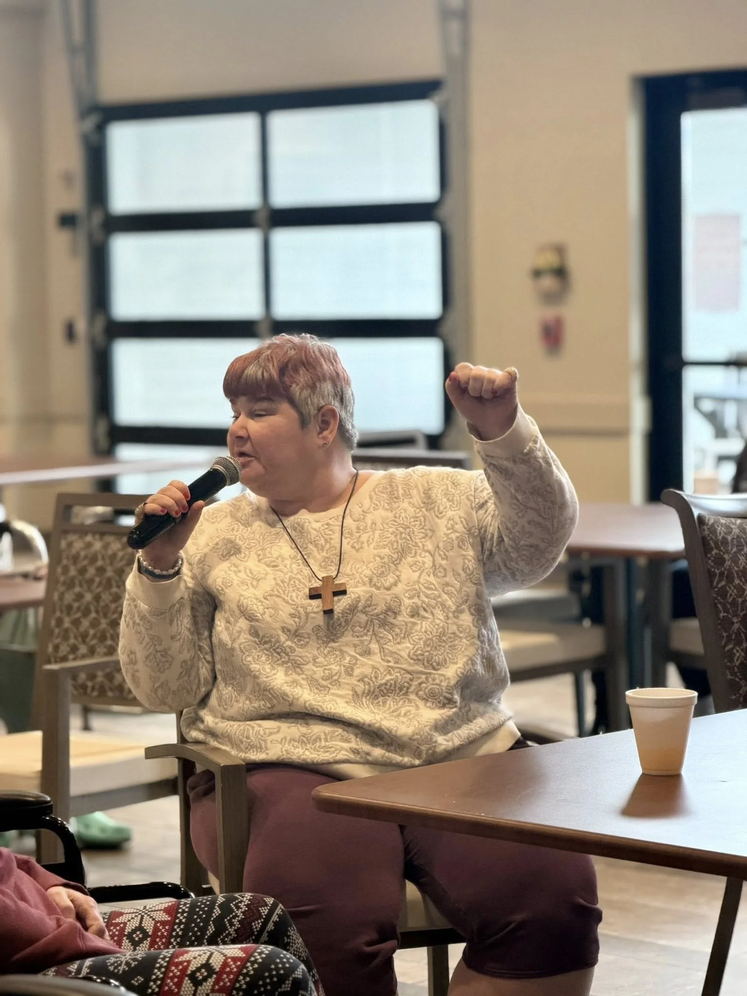 A woman with short, reddish hair and a cross necklace is sitting at a table, speaking into a microphone with her right hand raised in a fist. She is wearing a light-colored patterned top and maroon pants. There are chairs and tables in the background, and a large window with black frames.