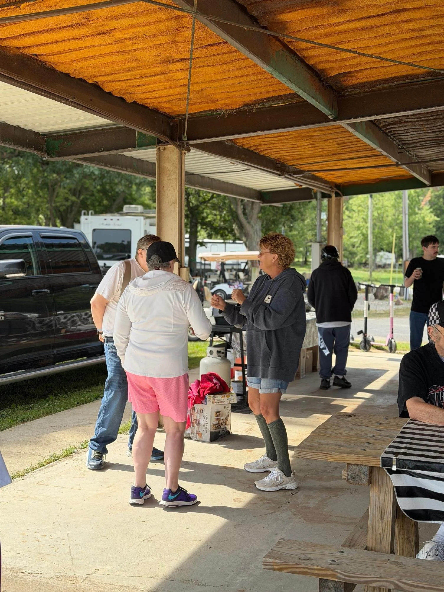 Group of people standing and talking under a wooden and metal shelter at an outdoor event, with vehicles and trees in the background.