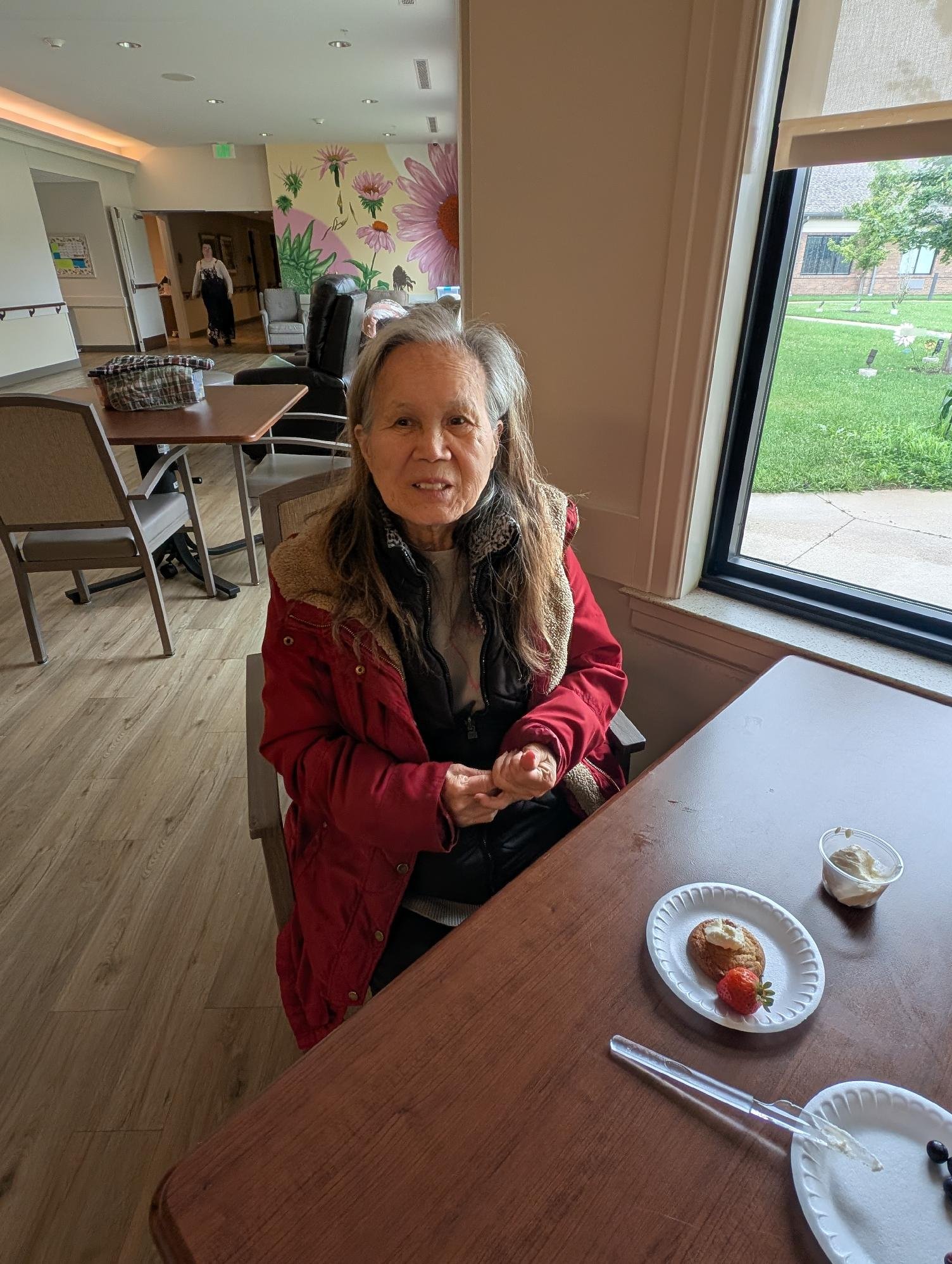 An elderly woman with gray hair seated at a table in a restaurant or cafe, wearing a red jacket, with a plate of desserts and a glass of whipped cream on the table. In the background, there is a colorful painted wall with flowers, a woman in black ap
