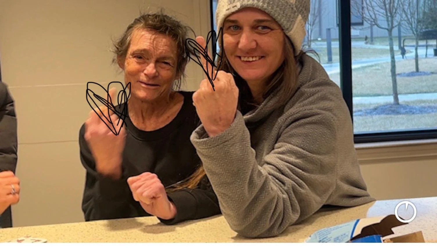 Two women smiling and arm wrestling at a table indoors, with a window showing trees and grass outside.