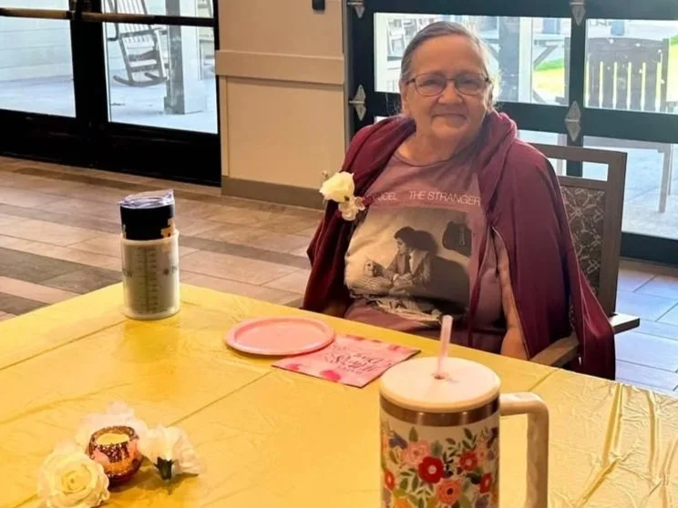 An elderly woman sitting at a table during daytime with a large mug, a pink plate, a pink napkin, a cupcake with a white flower, and a black container, in a room with large windows and a patio outside.