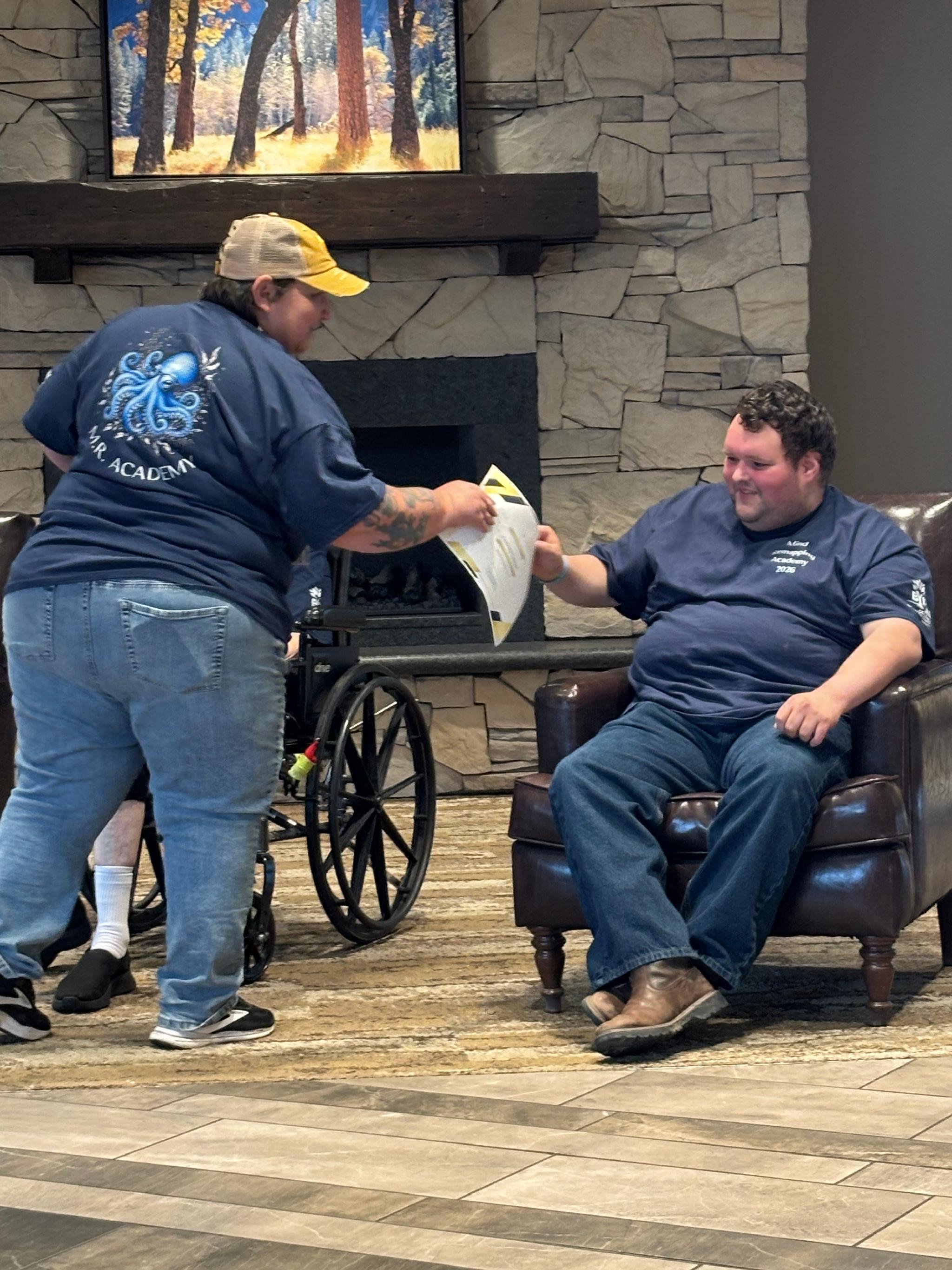 A man sitting in a leather armchair holding hands with a woman in a wheelchair. The man has curly hair, beard, and is wearing dark jeans and a navy shirt. The woman, standing next to him, has long dark hair, tattoos on her arm, and is wearing jeans, 