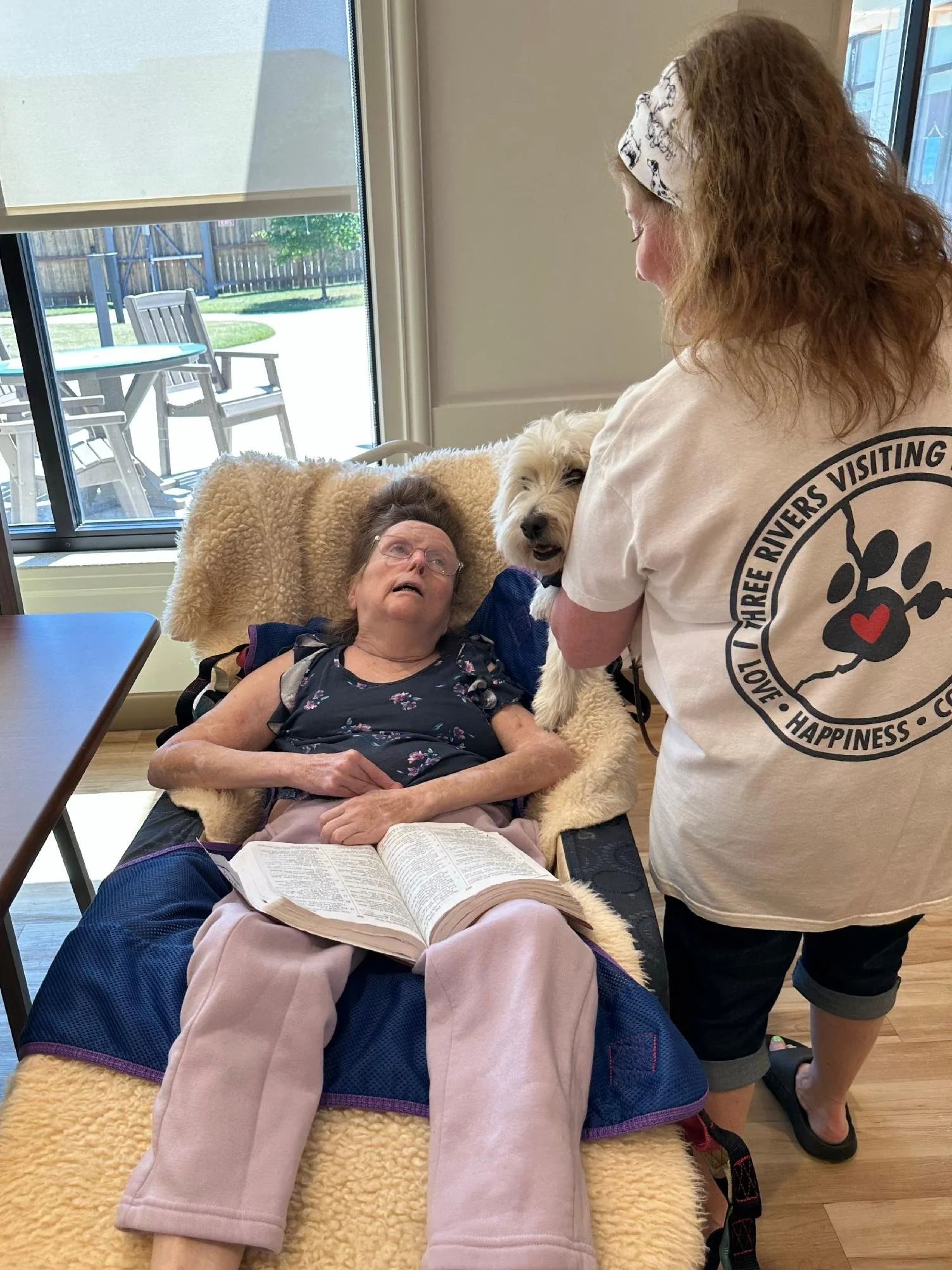 An elderly woman lying on a chair with a book, a woman wearing a shirt with a recentering paw print badge, and a dog with curly fur in an indoor setting near a window.