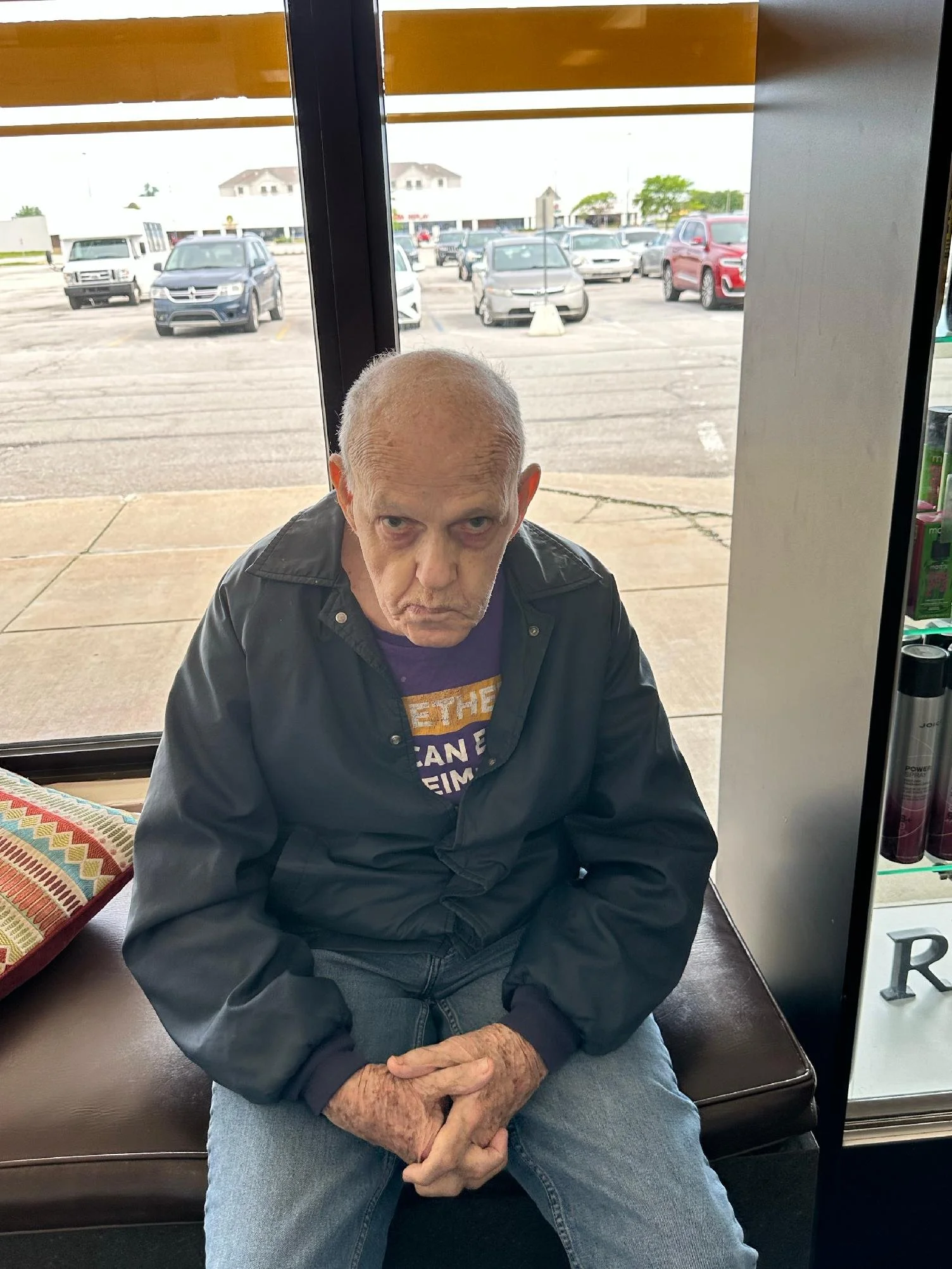 An elderly man with gray hair and wearing a black jacket and blue jeans is sitting on a bench inside a store, looking at the camera with a neutral expression. Behind him, there is a window showing a parking lot with several cars and a building in the