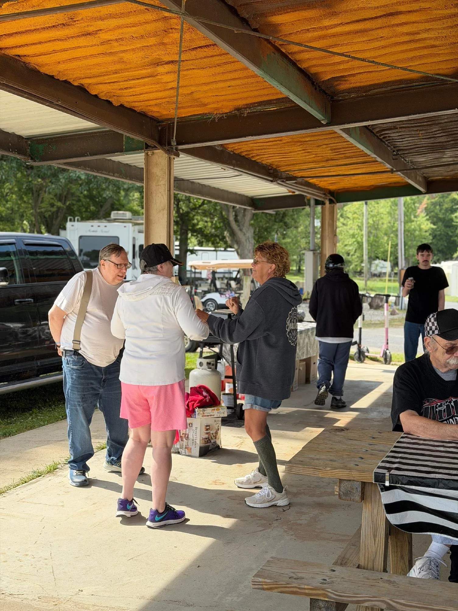 A group of people gathered under a pavilion outside, engaging in conversation, with trees and vehicles in the background.
