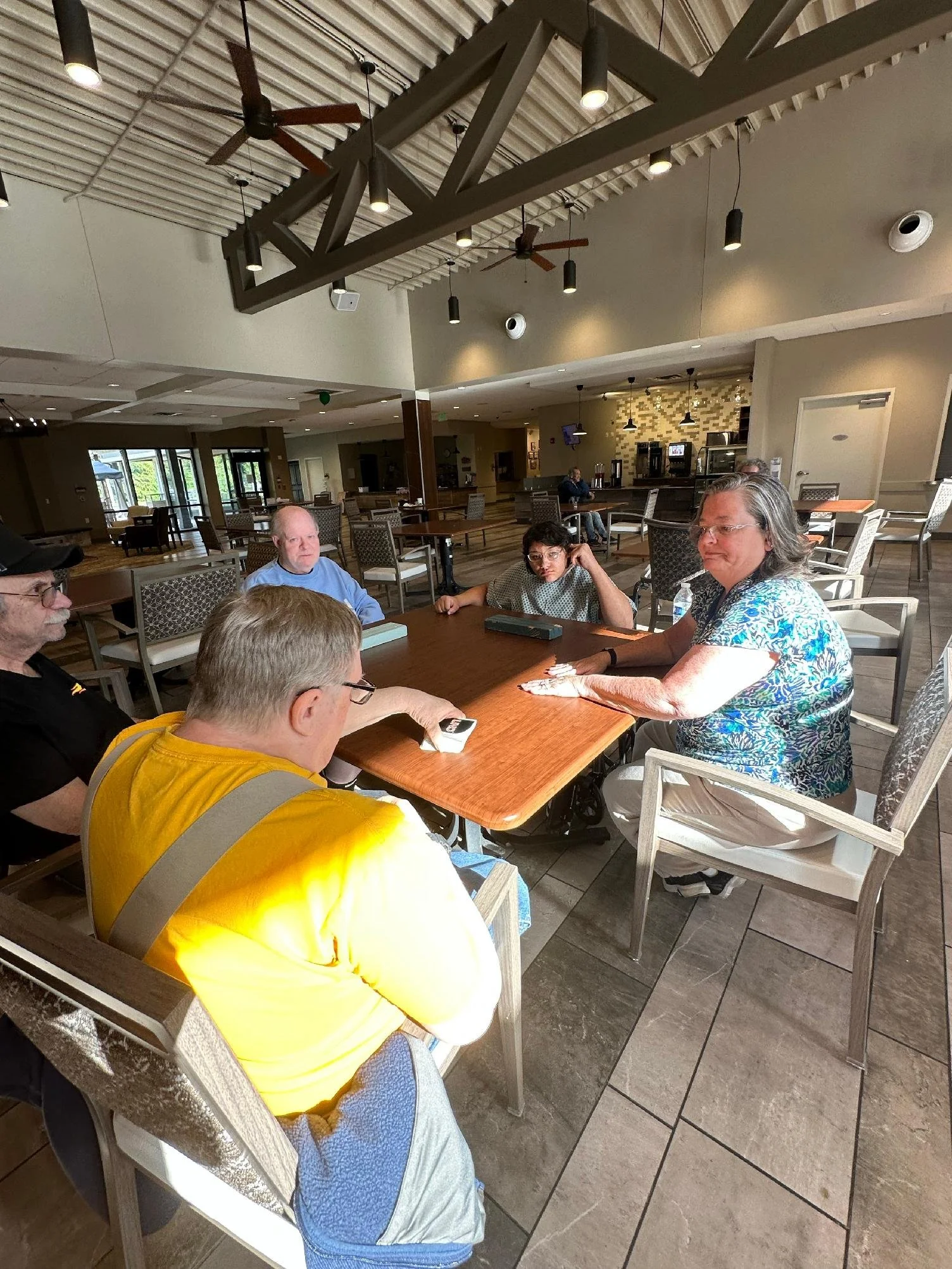 A group of people sitting around a rectangular table in a restaurant or cafe, with a bar area and large open windows in the background.