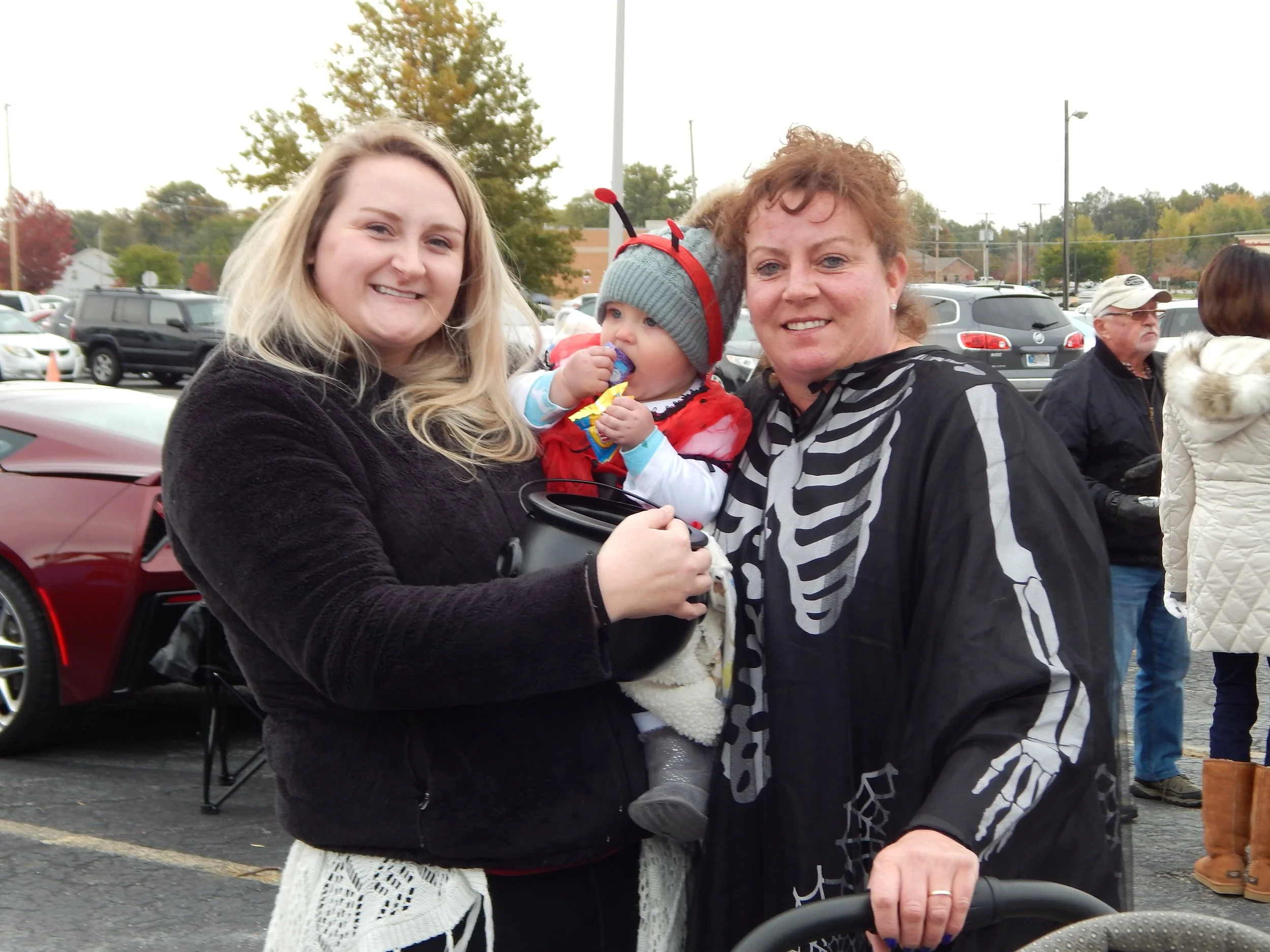 Photo of three people outdoors in a parking lot, smiling at the camera. Two women are holding a baby dressed in a red and gray costume with antennae, possibly a ladybug or similar insect. The background includes cars, trees with fall foliage, and other people.