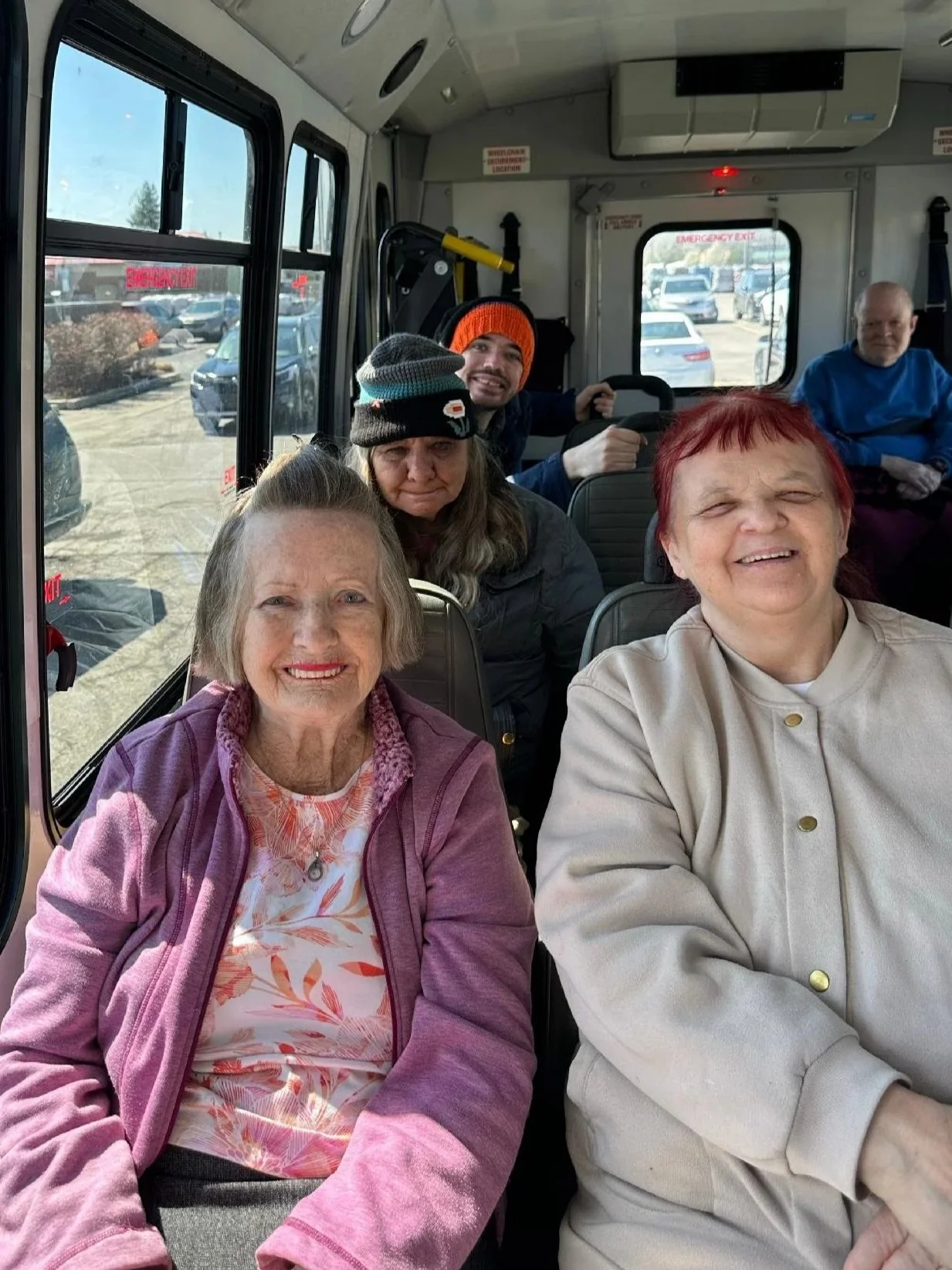 Inside a bus with six people, two older women in the foreground smiling at the camera, one with red hair and one with gray hair. Behind them, a woman with a hat and a man with a beanie are sitting, with other passengers visible in the background.