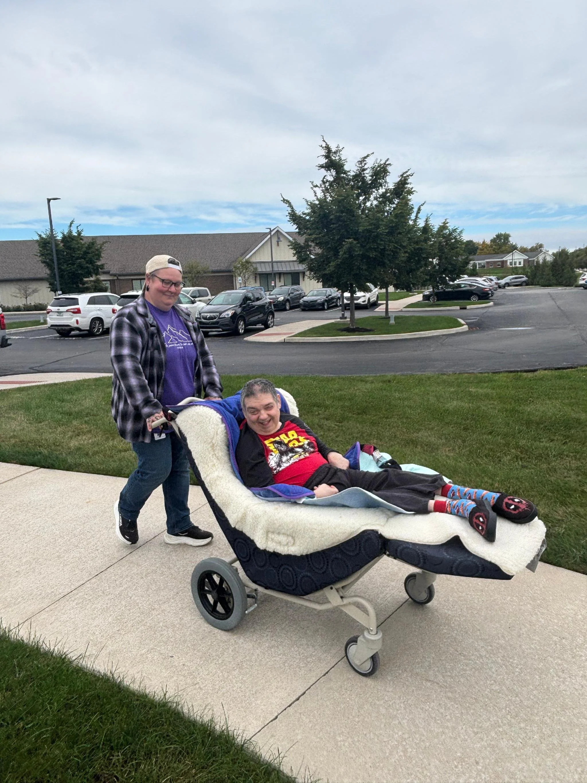 A person with glasses and a cap pushing a person in a stroller on a sidewalk in a parking lot with cars and trees in the background.