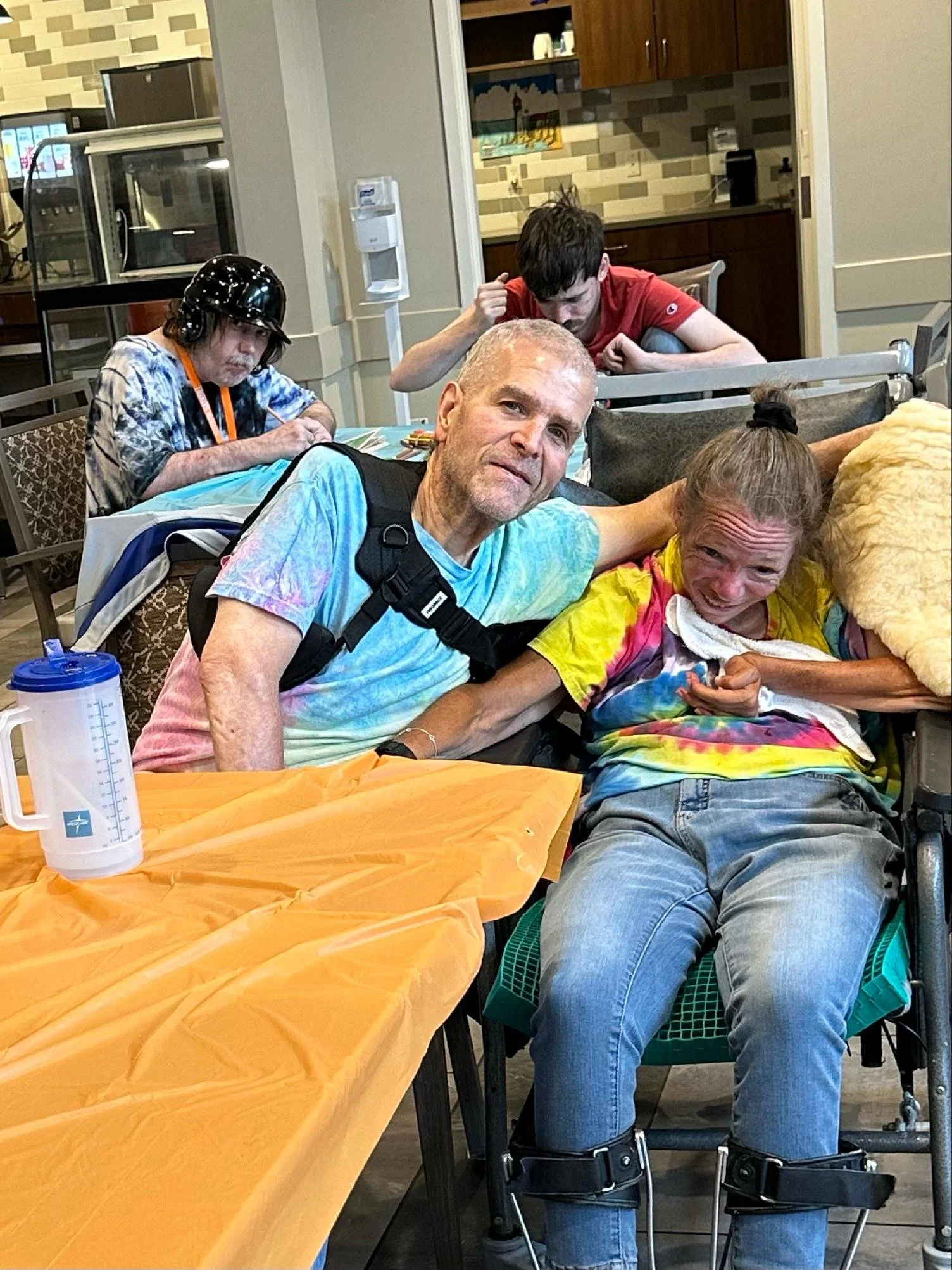 A group of people sitting at a table in a dining area, including a woman with a ladybug costume, an older man with prosthetic legs, and a woman with a colorful tie-dye shirt. The background shows a kitchen with wooden cabinets.