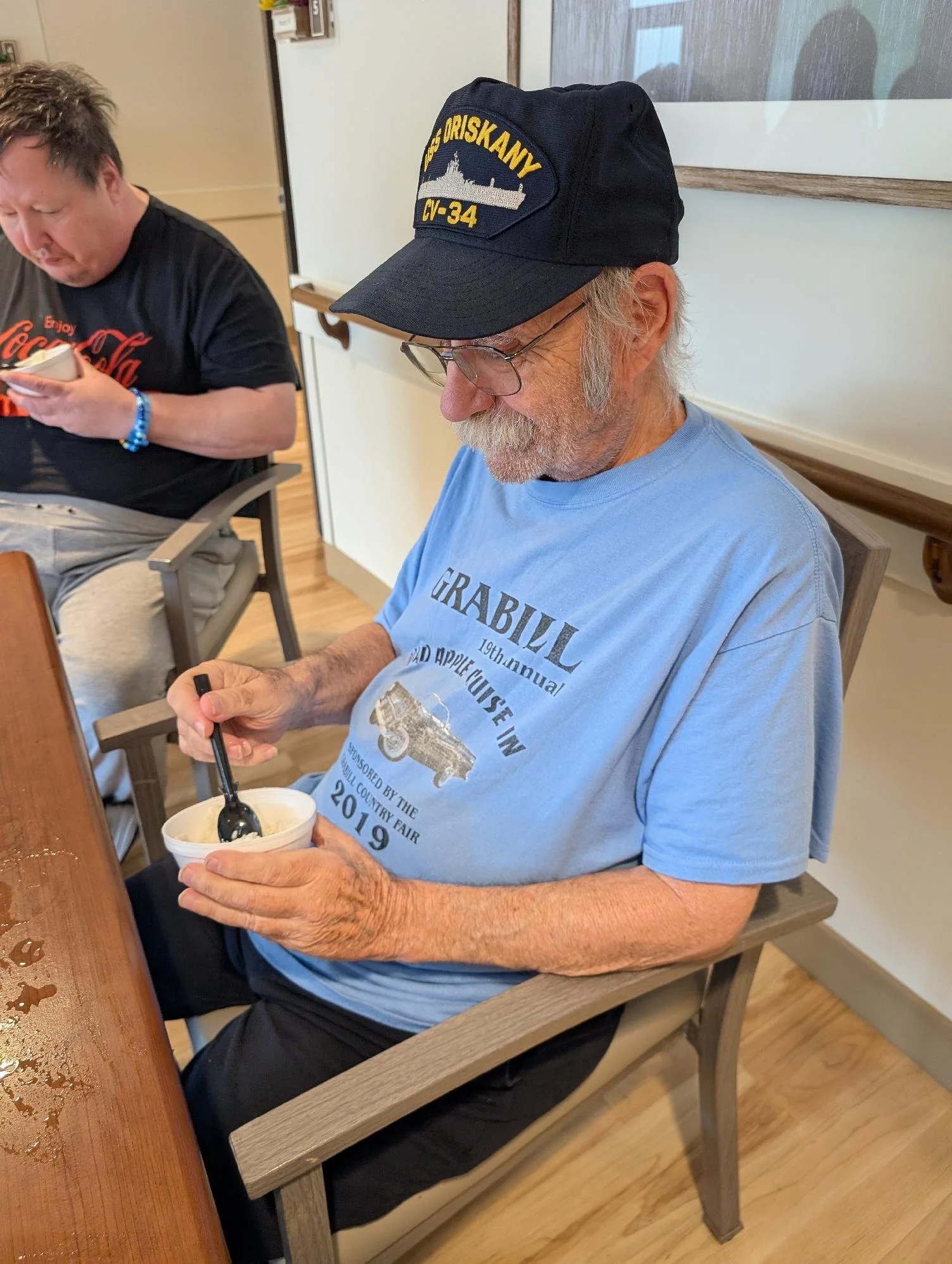 An elderly man with gray hair, glasses, and a beard wearing a black cap and a blue T-shirt, sitting on a wooden chair, holding a bowl of ice cream and eating it with a spoon. There is a woman next to him wearing a black T-shirt with orange text and a