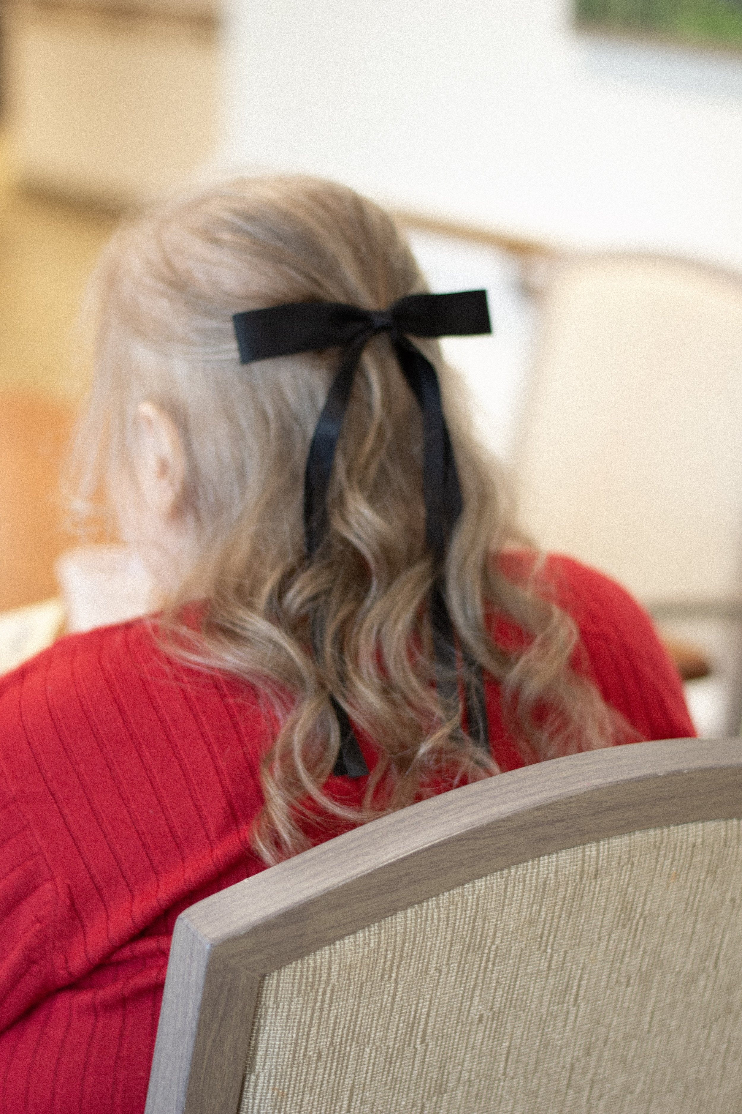 Back of a woman with long blond hair wearing a black bow and a red sweater, sitting at a desk or table, with a beige upholstered backrest in front of her.