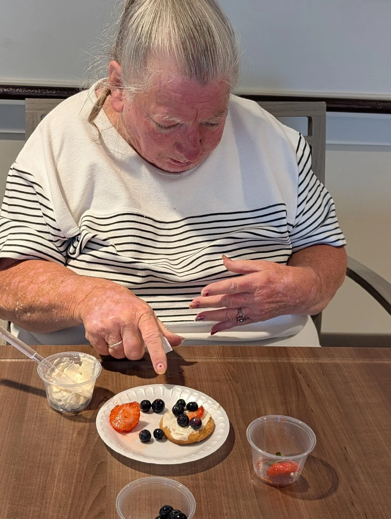 An elderly woman with white hair and a striped shirt is sitting at a wooden table, pointing at a plate of assorted desserts topped with fruits, with small cups of additional desserts or drinks around her.