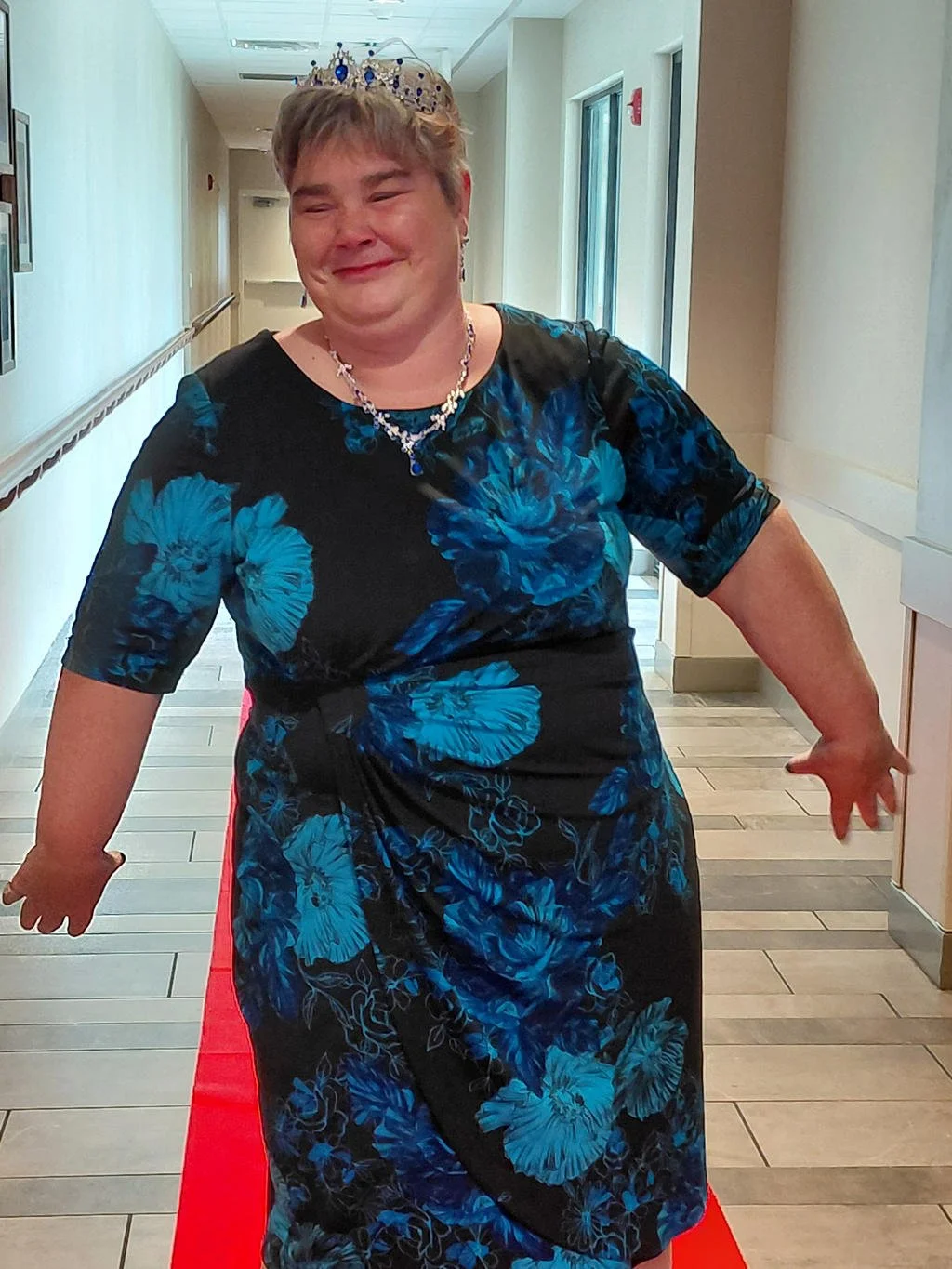 A woman wearing a crown, necklace, and earrings is standing on a red carpet in a hallway, smiling and posing for the camera.