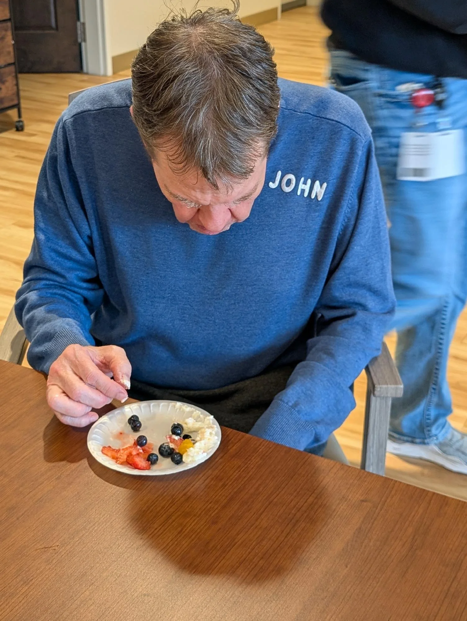An elderly man wearing a blue sweatshirt with the name tag 'JOHN' sitting at a wooden table, looking down at a paper plate with strawberries, blueberries, and whipped cream.