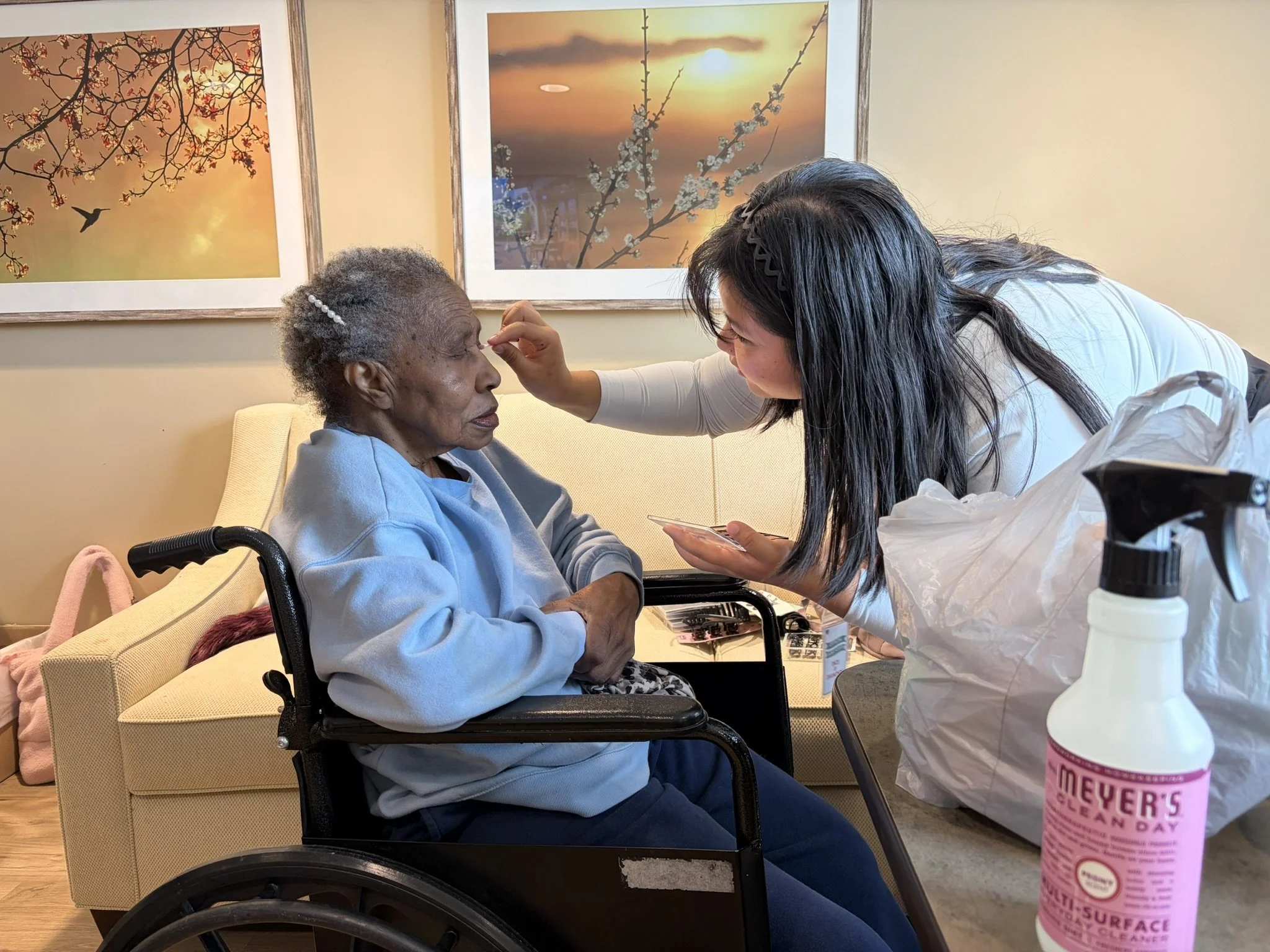 A young woman with dark hair and a black hair clip is applying makeup to an elderly woman with gray hair in a blue sweatshirt, seated in a wheelchair. They are in a room decorated with framed floral and sunset artwork on the wall.