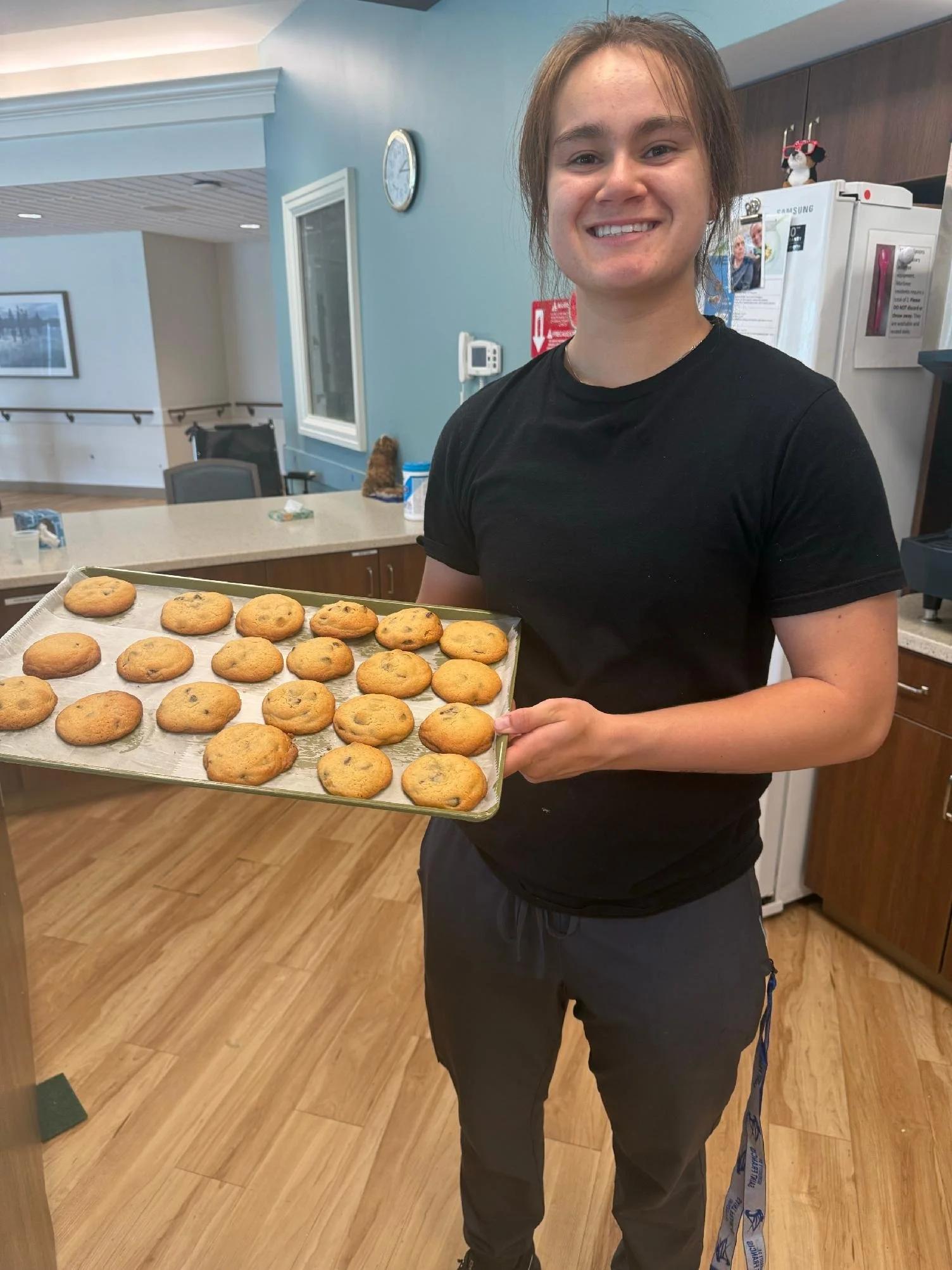 A smiling young woman holding a tray of freshly baked cookies in a kitchen or cafe setting.