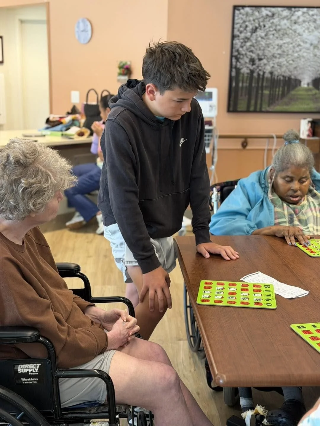 A young boy standing next to a table with Bingo cards, assisting two elderly women who are playing Bingo, one in a wheelchair and the other sitting in a chair, in a room with warm-colored walls and a large wall art of a white flowering tree.