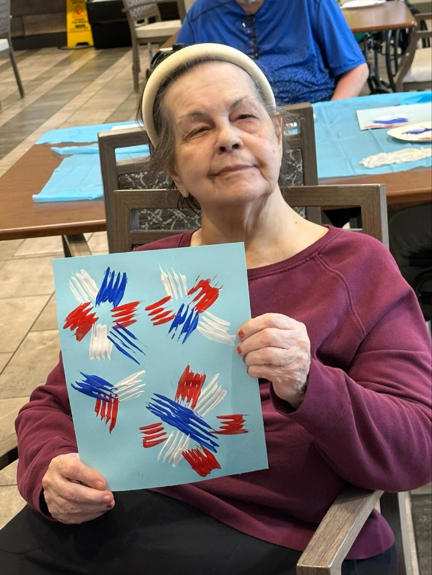 An elderly woman wearing a beige beanie and maroon sweater sitting at a wooden table in a restaurant, holding a colorful abstract painting with red, blue, and white brushstrokes.