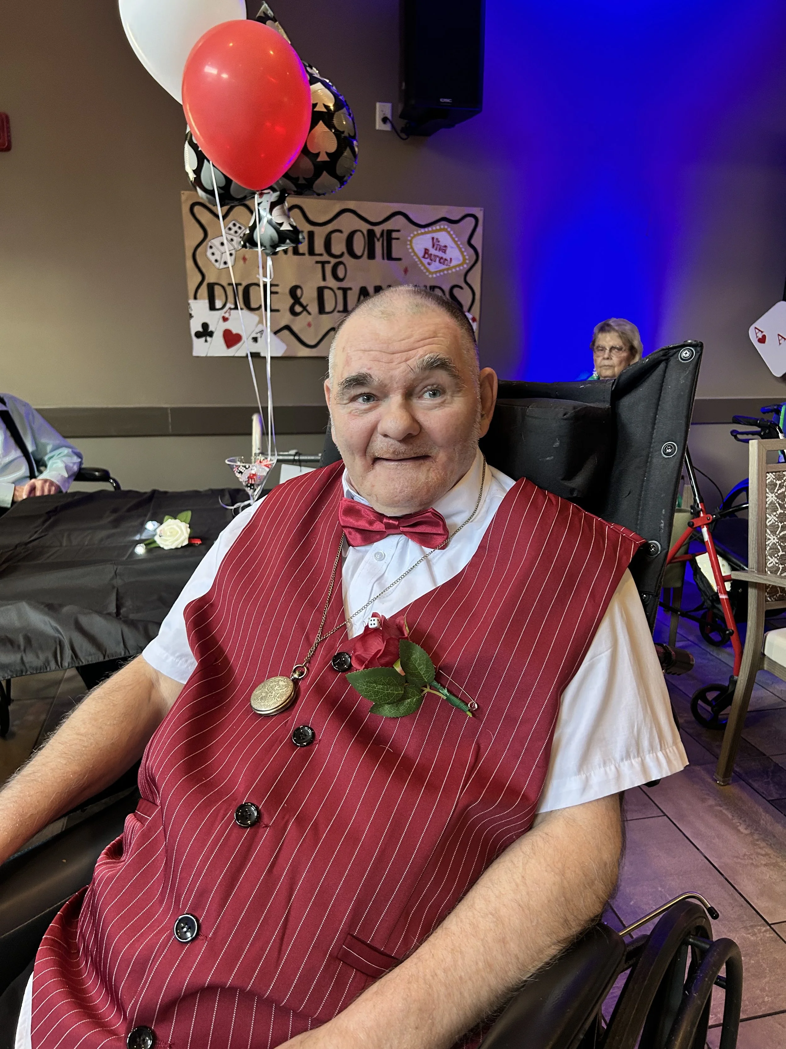 A man wearing a red pinstripe vest and bow tie, sitting in a wheelchair, with a boutonniere on his vest, and balloons in the background at a celebration event.
