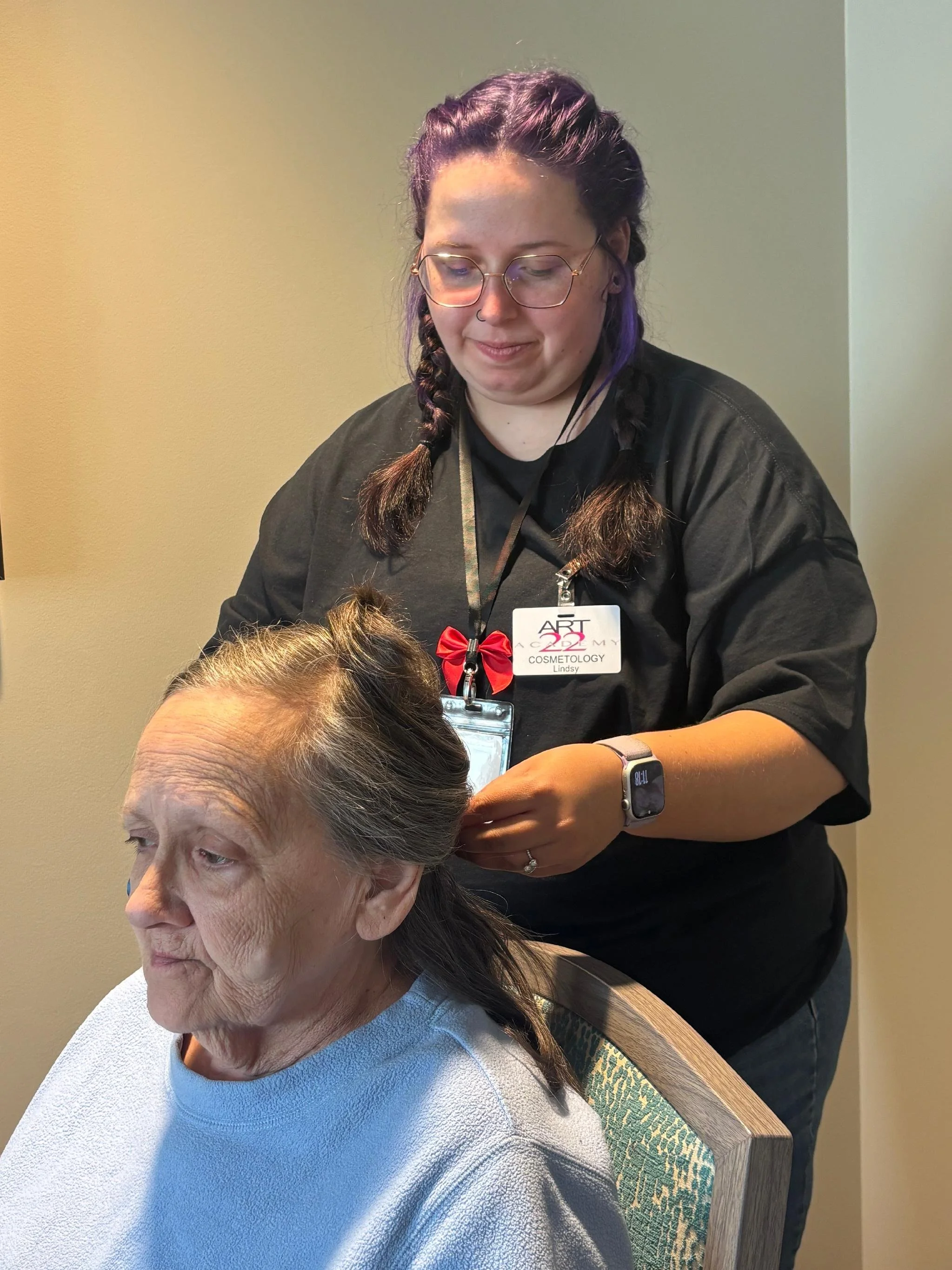 A young woman with purple hair in braids and glasses is providing a cosmetic service to an older woman sitting in a chair. The technician wears a black shirt, a lanyard with a name badge, and a smartwatch. She is smiling slightly as she works on the 