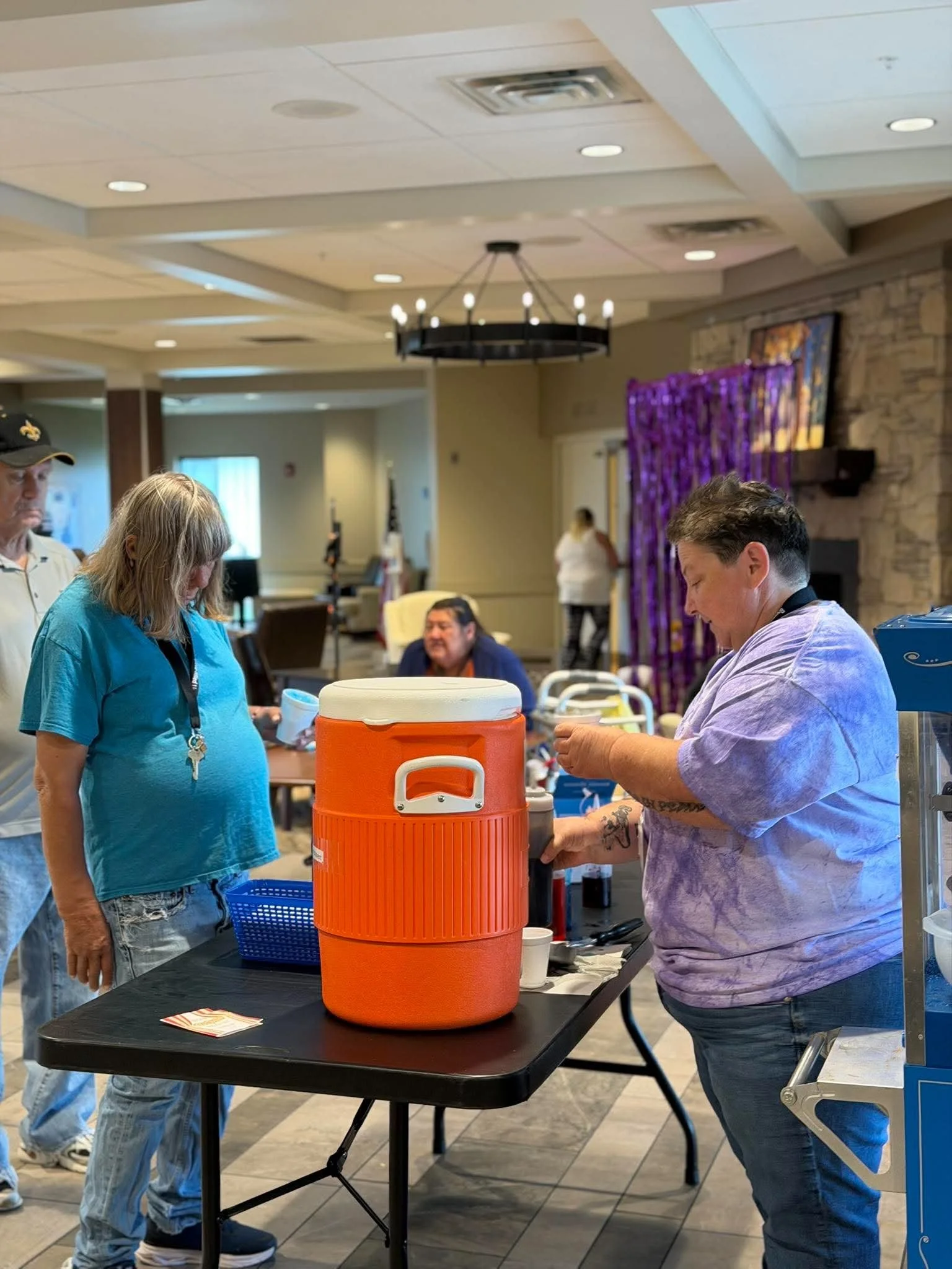 People at a reception or event table, with a large orange and white cooler in the center. Several people are engaged in conversation or receiving items, with a woman in a purple shirt and a woman in a turquoise shirt visible. The background includes 