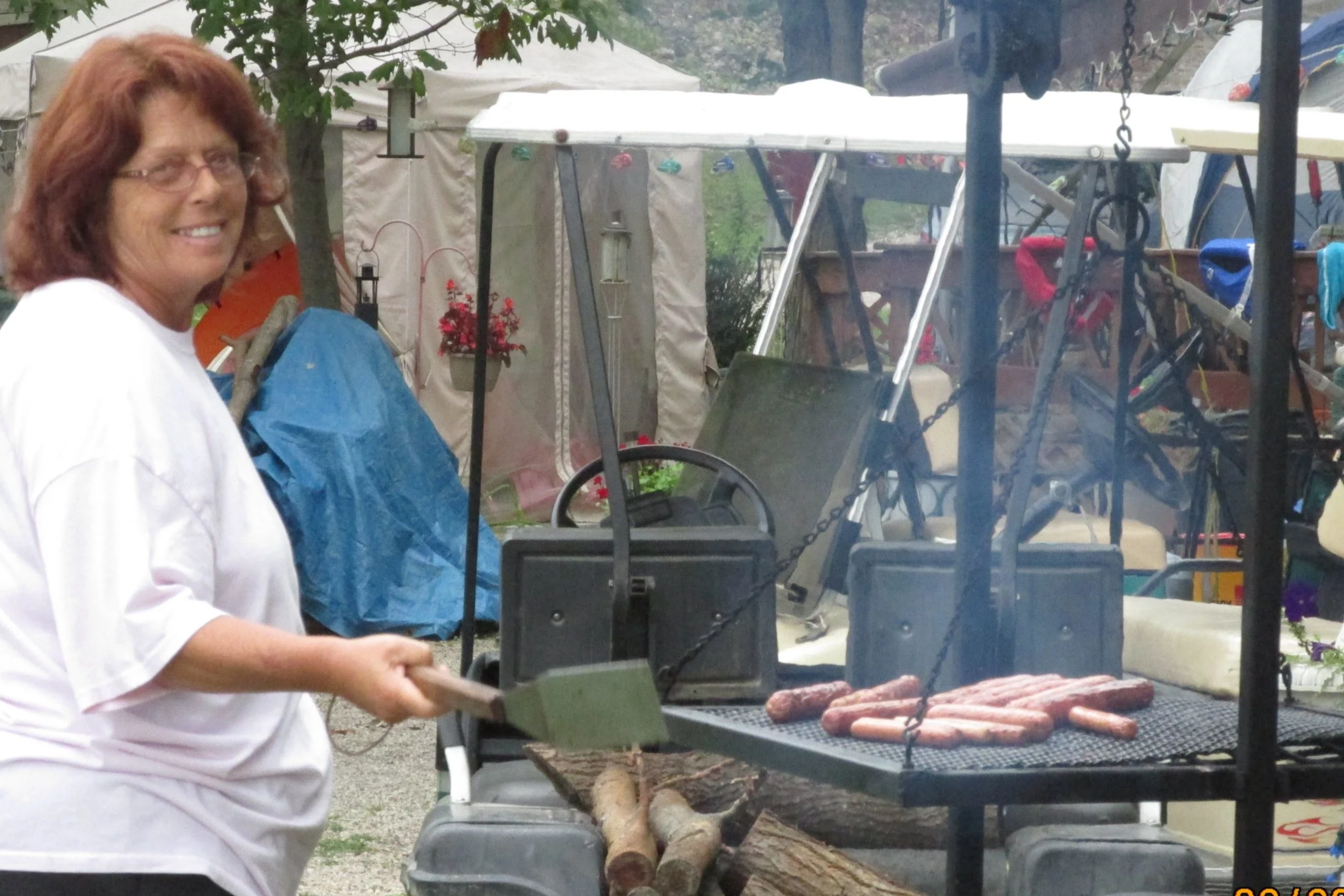 A woman with red hair wearing glasses and a white shirt is grilling hot dogs and sausages on an outdoor barbecue. She is smiling and standing near the grill, which is filled with cooked meats. In the background, there are trees, tents, and various camping or outdoor equipment.