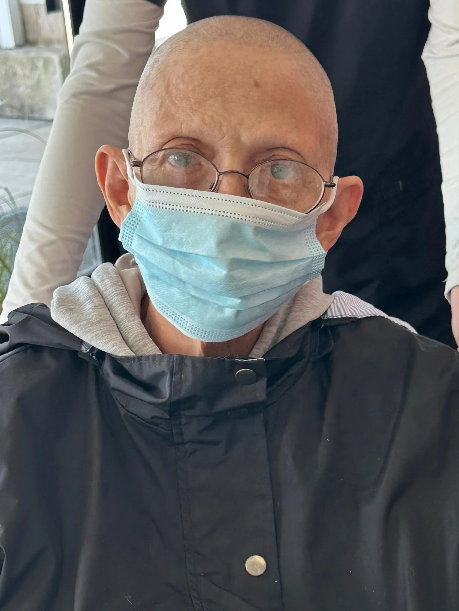 Close-up of a person wearing glasses and a surgical mask, dressed in a black jacket and a light-colored hoodie inside a building.