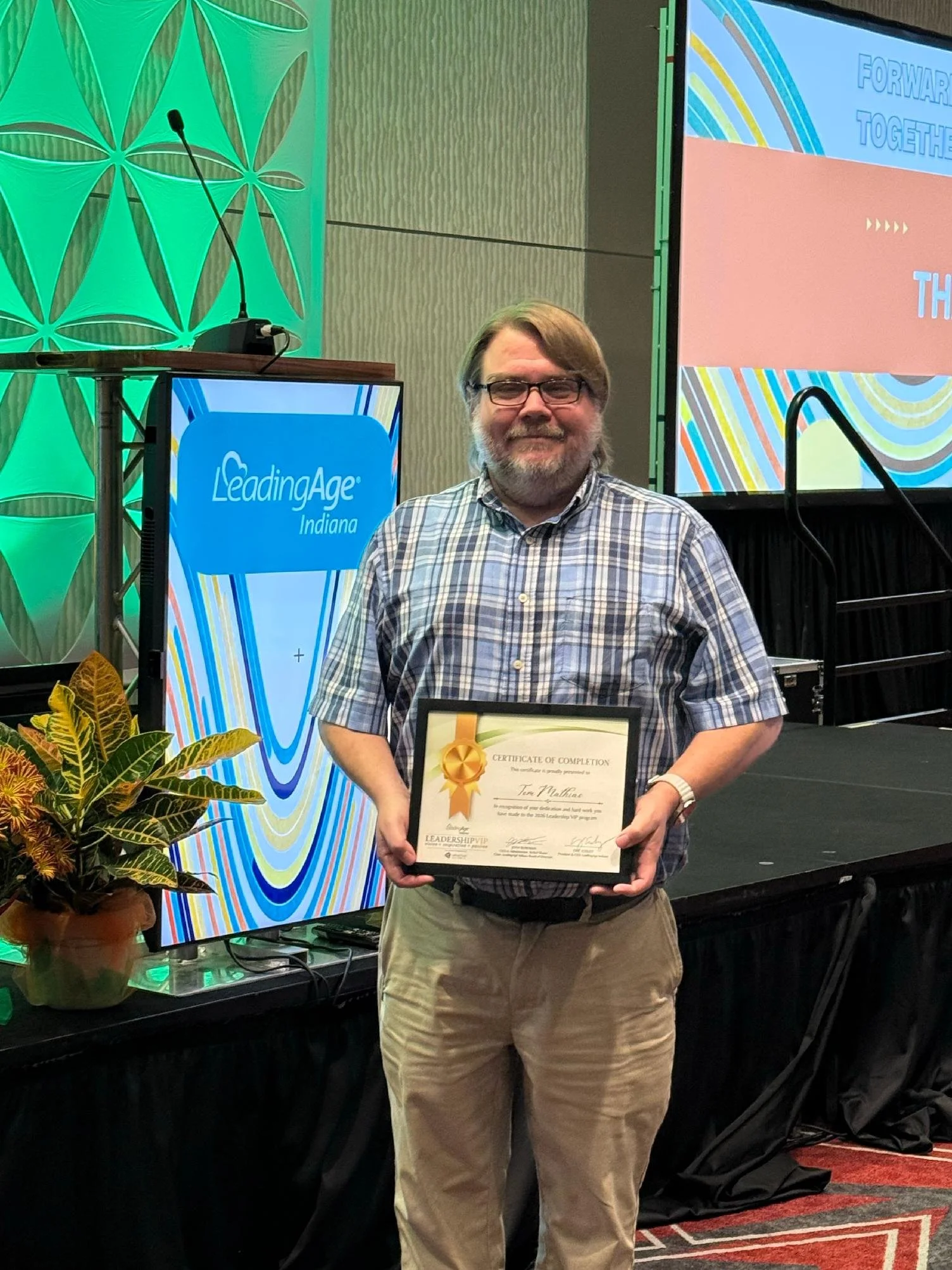 A man wearing glasses and a plaid shirt holding a framed certificate of completion at an indoor event with colorful displays and a podium in the background.