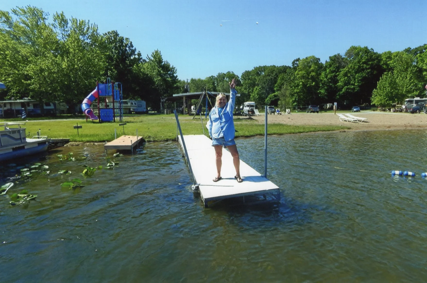 A woman standing on a floating dock at a lake beach, waving, with trees, a playground, and parked cars in the background during daytime.