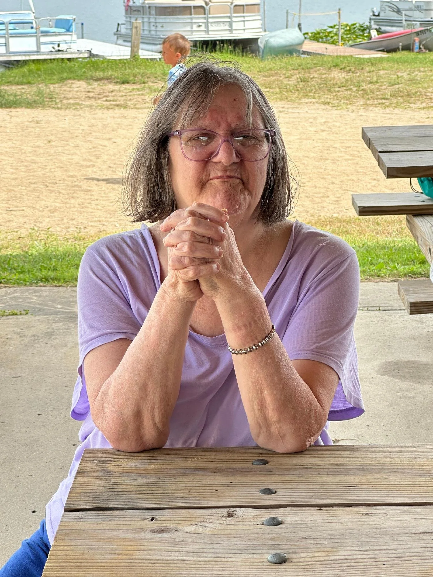 An elderly woman with gray hair and glasses sitting at a wooden table outdoors, with hands clasped in front of her face, looking directly at the camera. In the background, a young boy and boats on a waterway are visible.