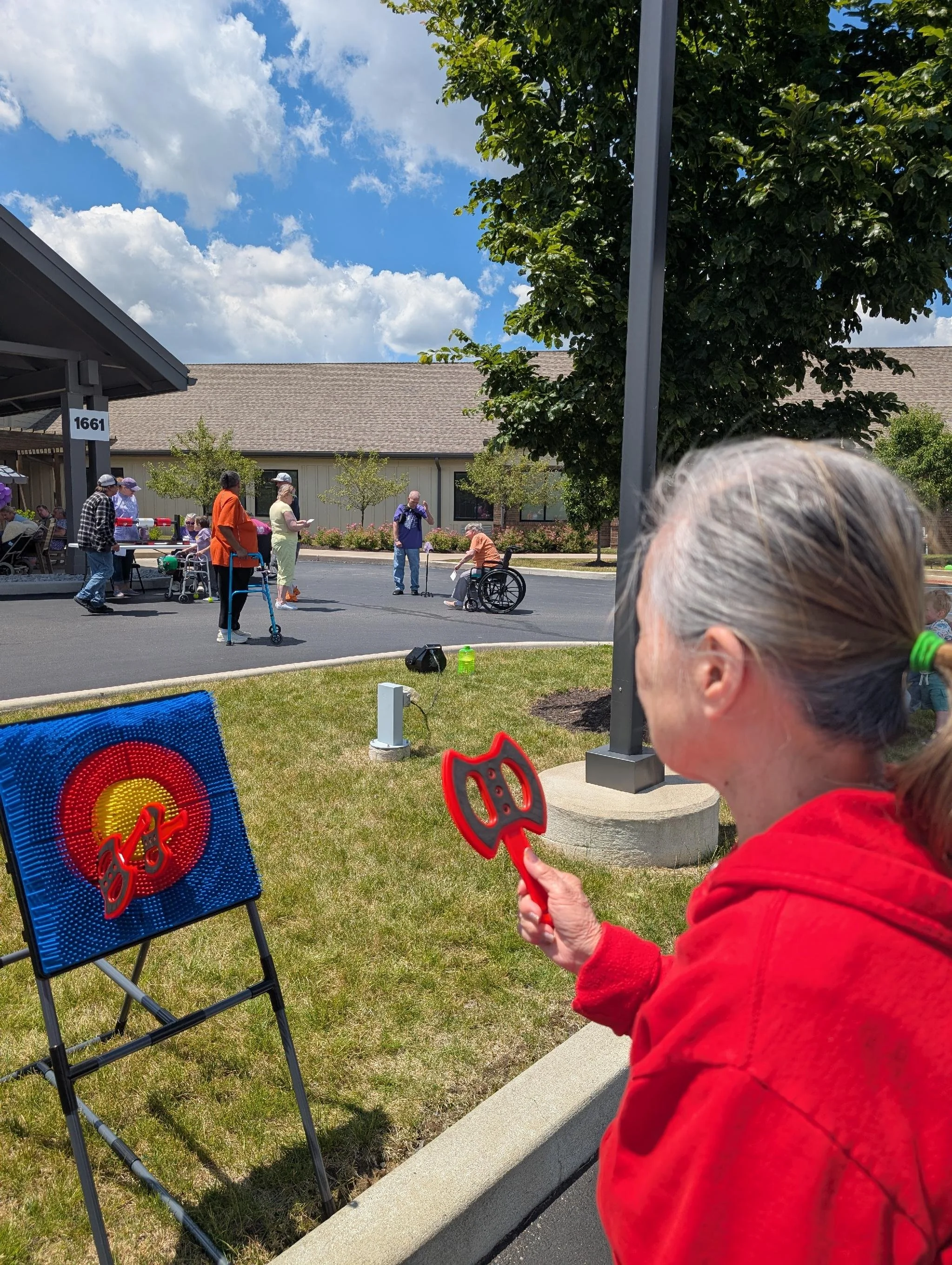 A woman in a red jacket is playing a target game outdoors, aiming a red toy gun at a police-themed target on a folding chair. Several people are gathered in the background, some standing and some sitting, near a building with a sign numbered 1661 und