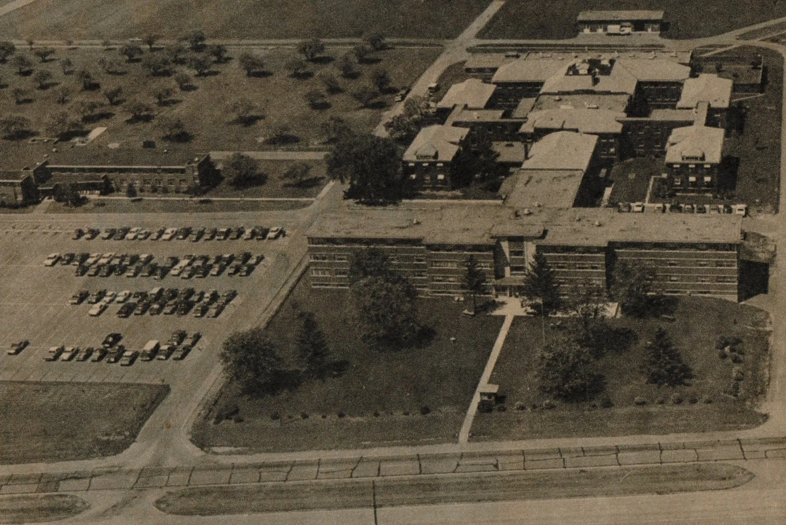 An aerial view of a school campus with multiple buildings, a large parking lot filled with cars, and surrounding green areas with trees.