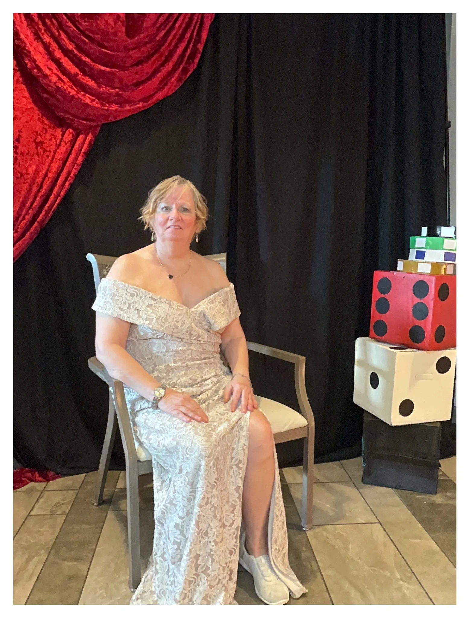 A woman in a white lace dress sitting on a beige chair, with one leg exposed and wearing a white sneaker. The background features black curtains with red velvet draping on the left side and colorful large dice stacked on a side table on the right.