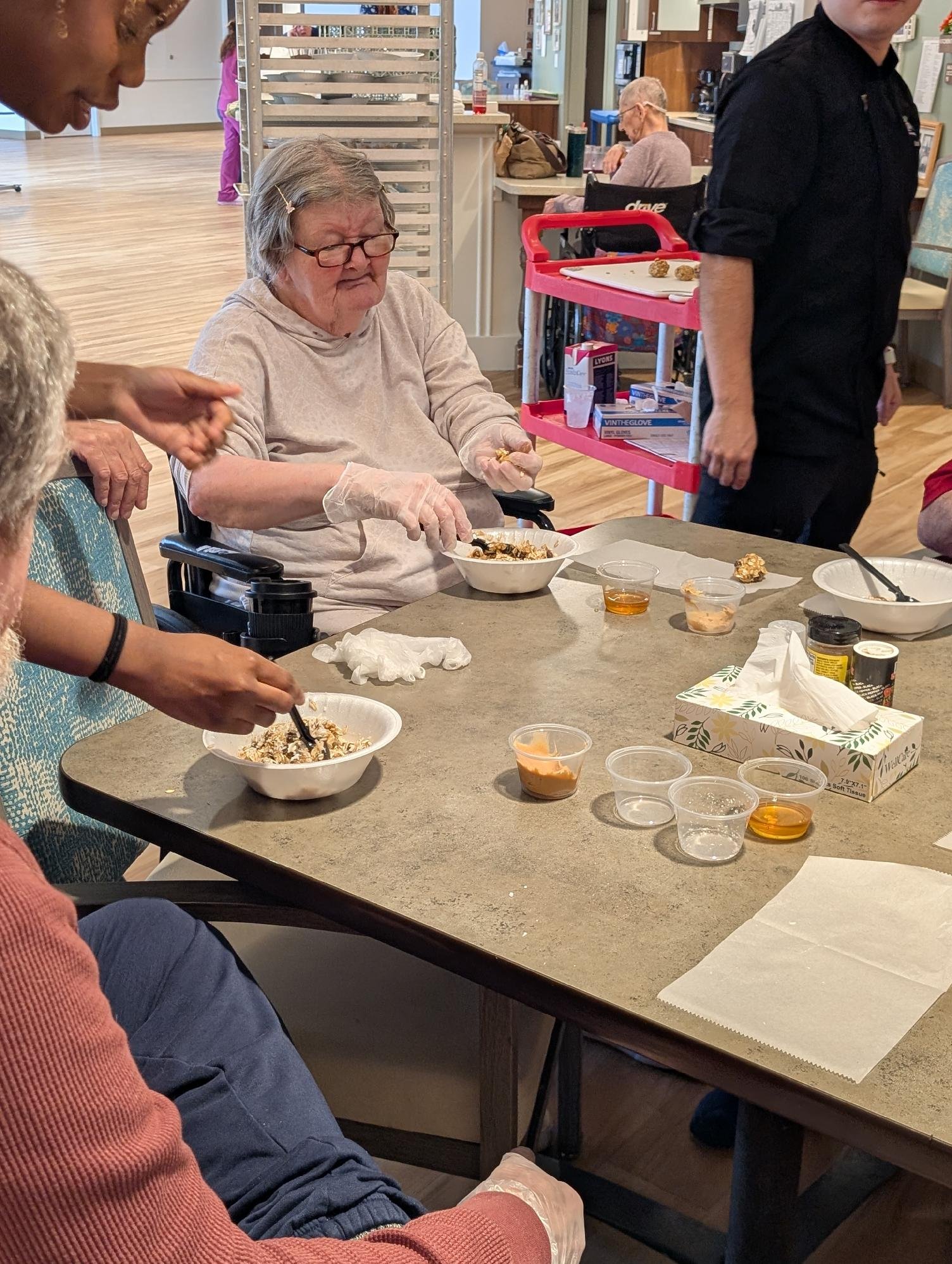 A group of elderly people sitting and standing around a table in a nursing home or assisted living facility, with one woman in a wheelchair serving herself food and a staff member pushing a cart in the background.