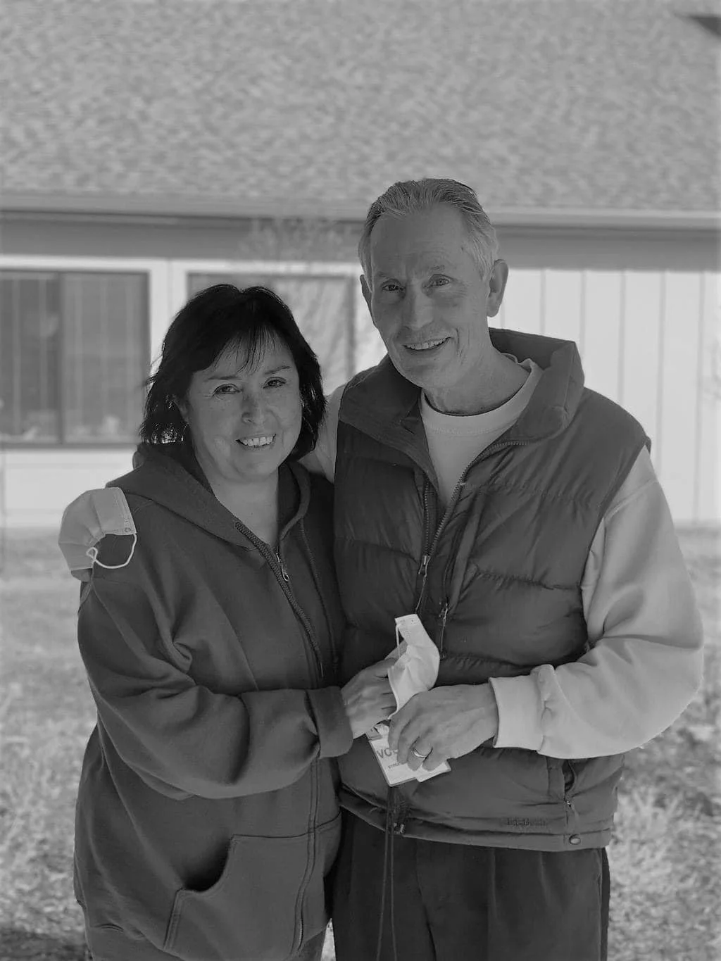 A grayscale photo of a woman and a man embracing outdoors in front of a house, smiling at the camera.